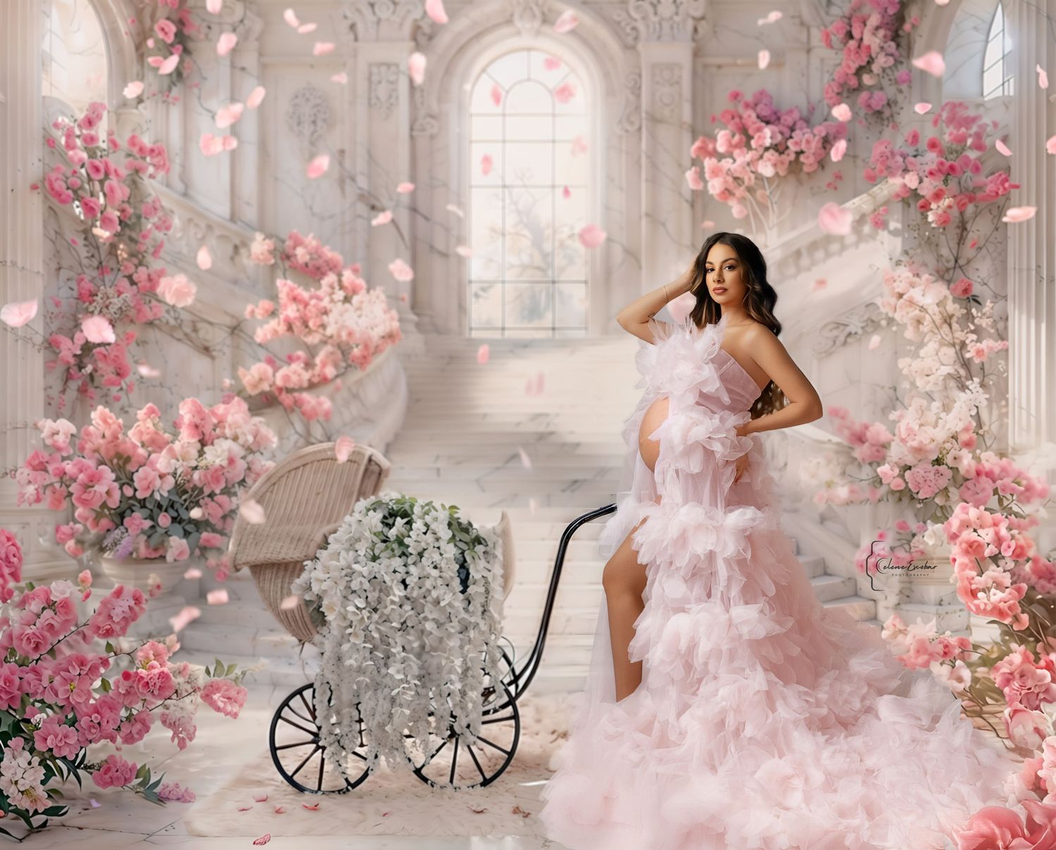 Pregnant woman in pink ruffle gown poses in elegant floral studio with white baby carriage and pink blossoms.