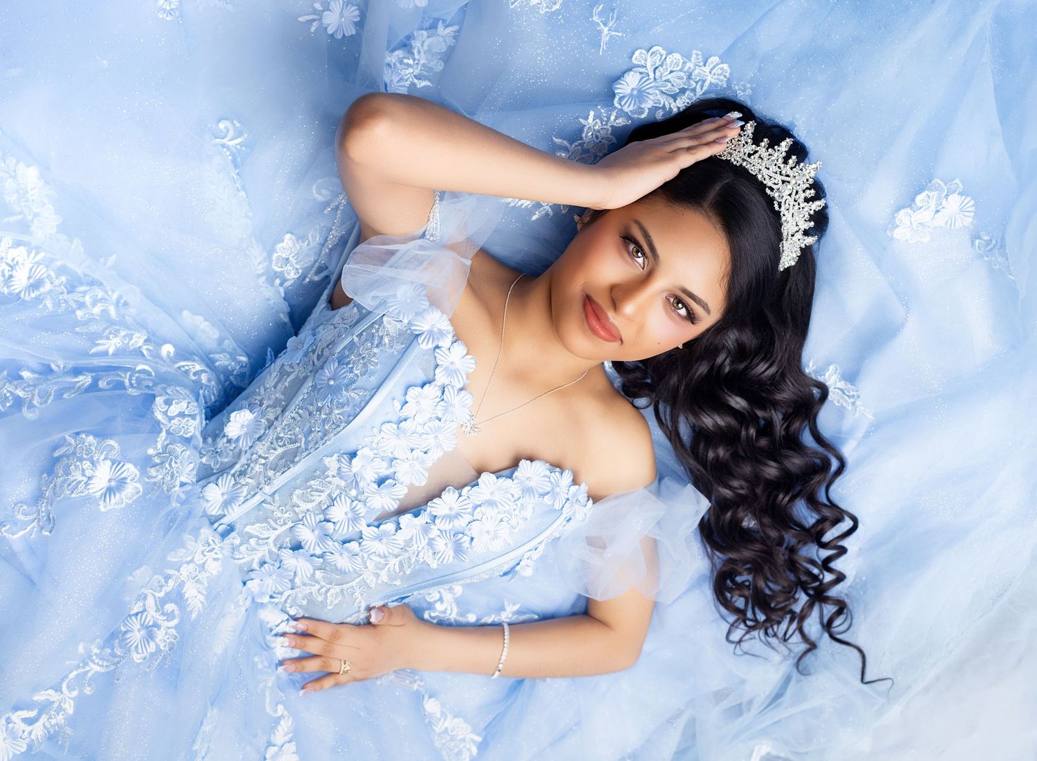 A young woman in a light blue off-shoulder ball gown with lace details and a silver tiara poses elegantly.