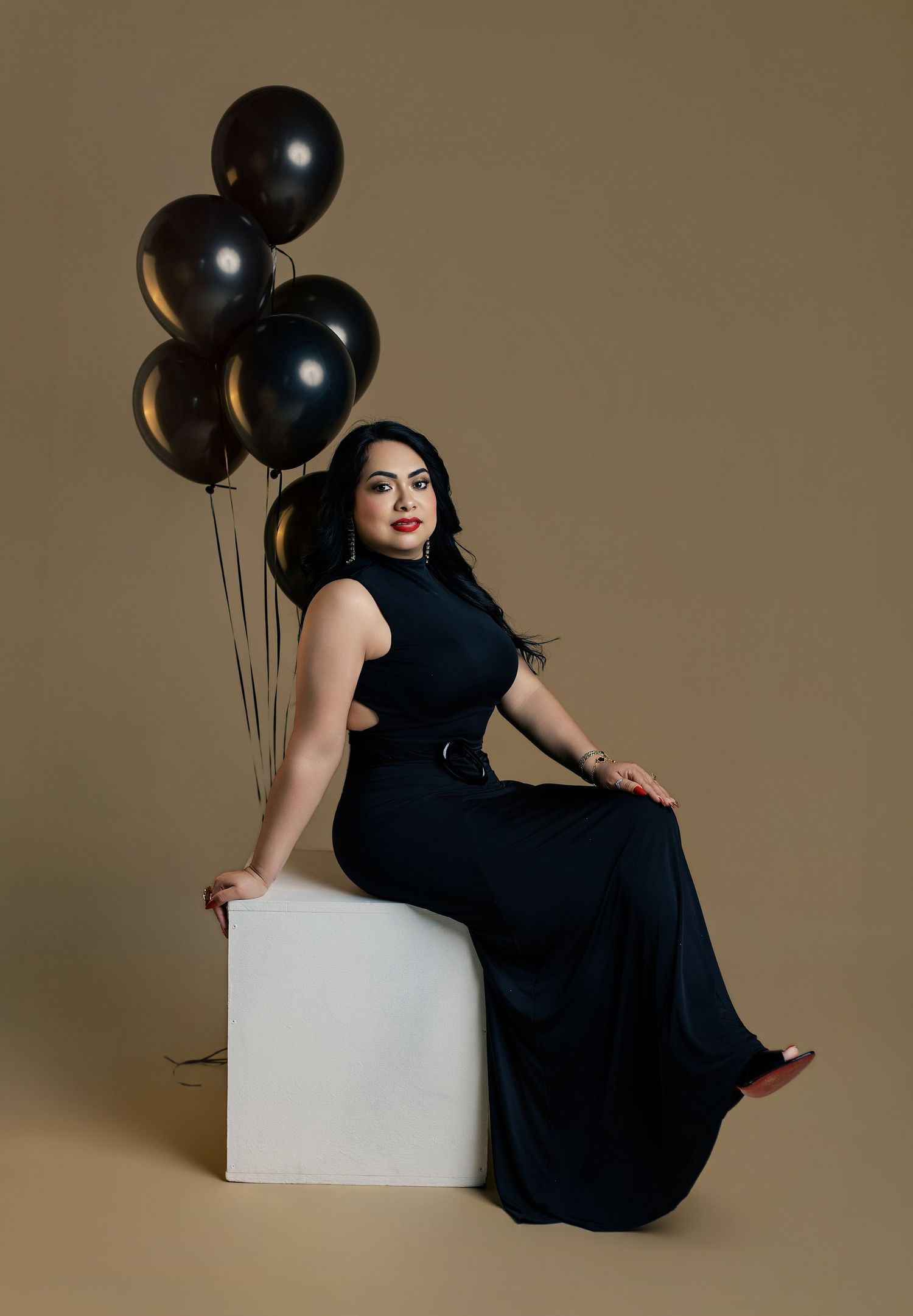 Woman in black gown seated on white cube with black balloons on tan background.