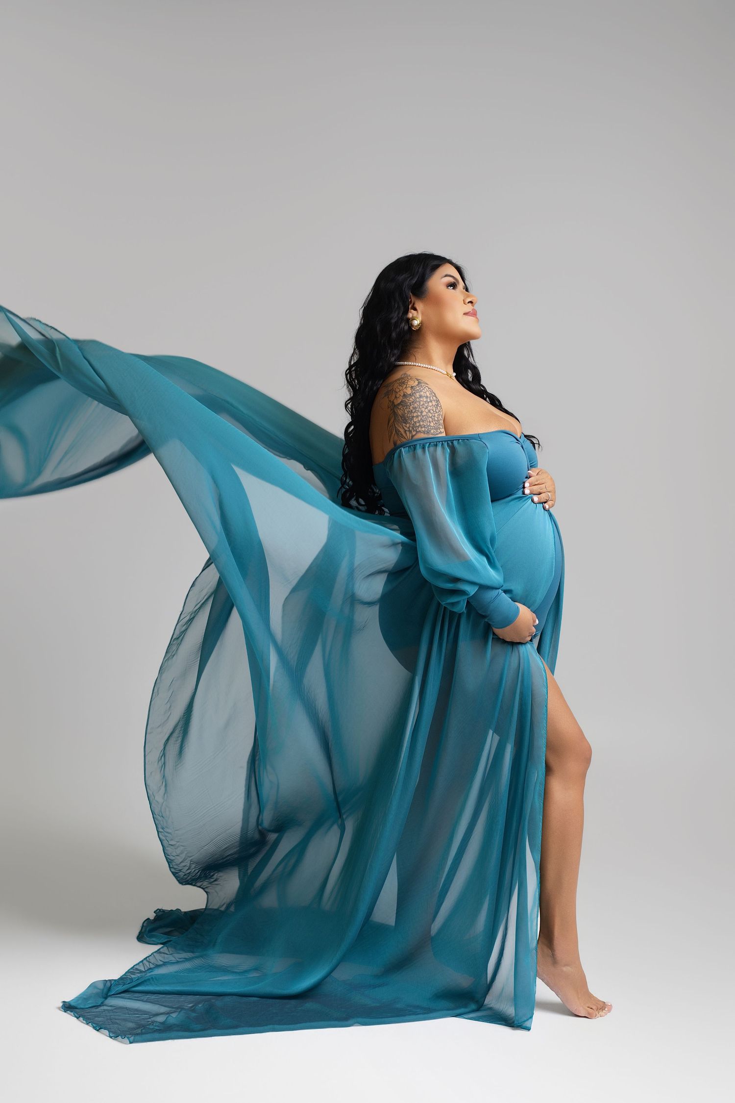 Pregnant woman in flowing teal chiffon gown cradling baby bump, looking up gracefully against gray background.