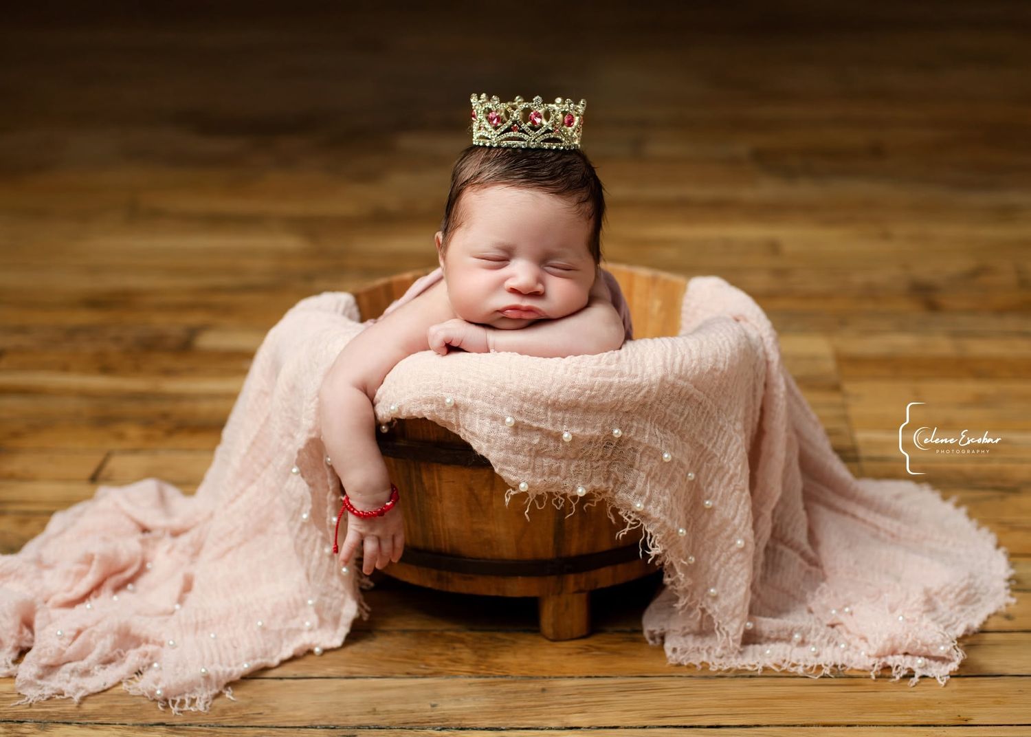 Newborn baby sleeping in wooden bowl wearing tiny crown with pink lace wrap, professional photography.