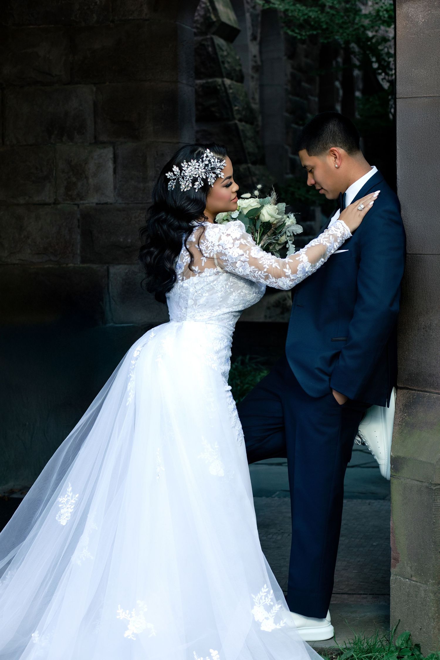 Bride in white lace gown with floral headpiece and groom in navy suit share romantic moment by stone wall.
