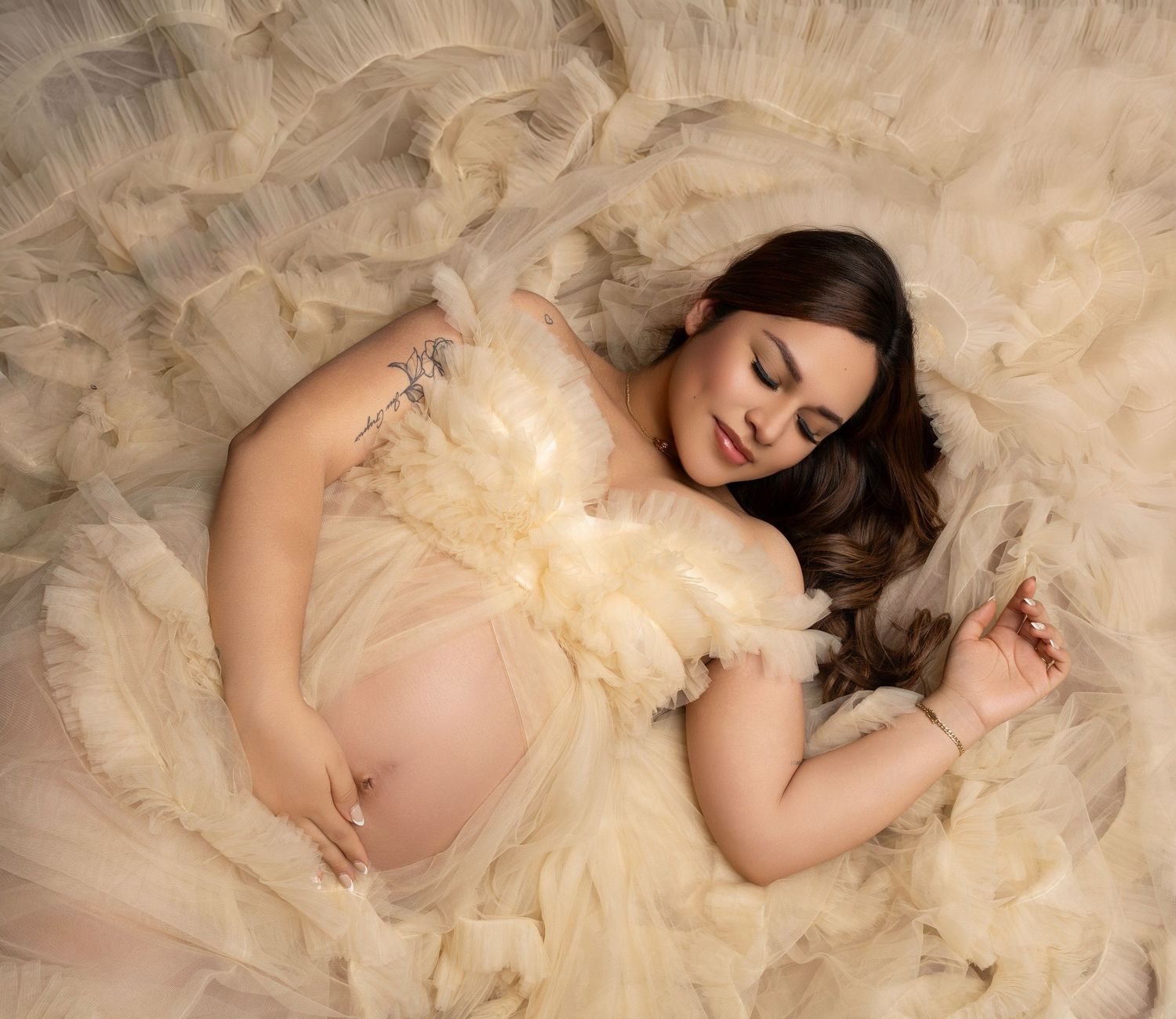 Pregnant woman in cream tulle gown lying gracefully, maternity photography session with soft elegant styling.