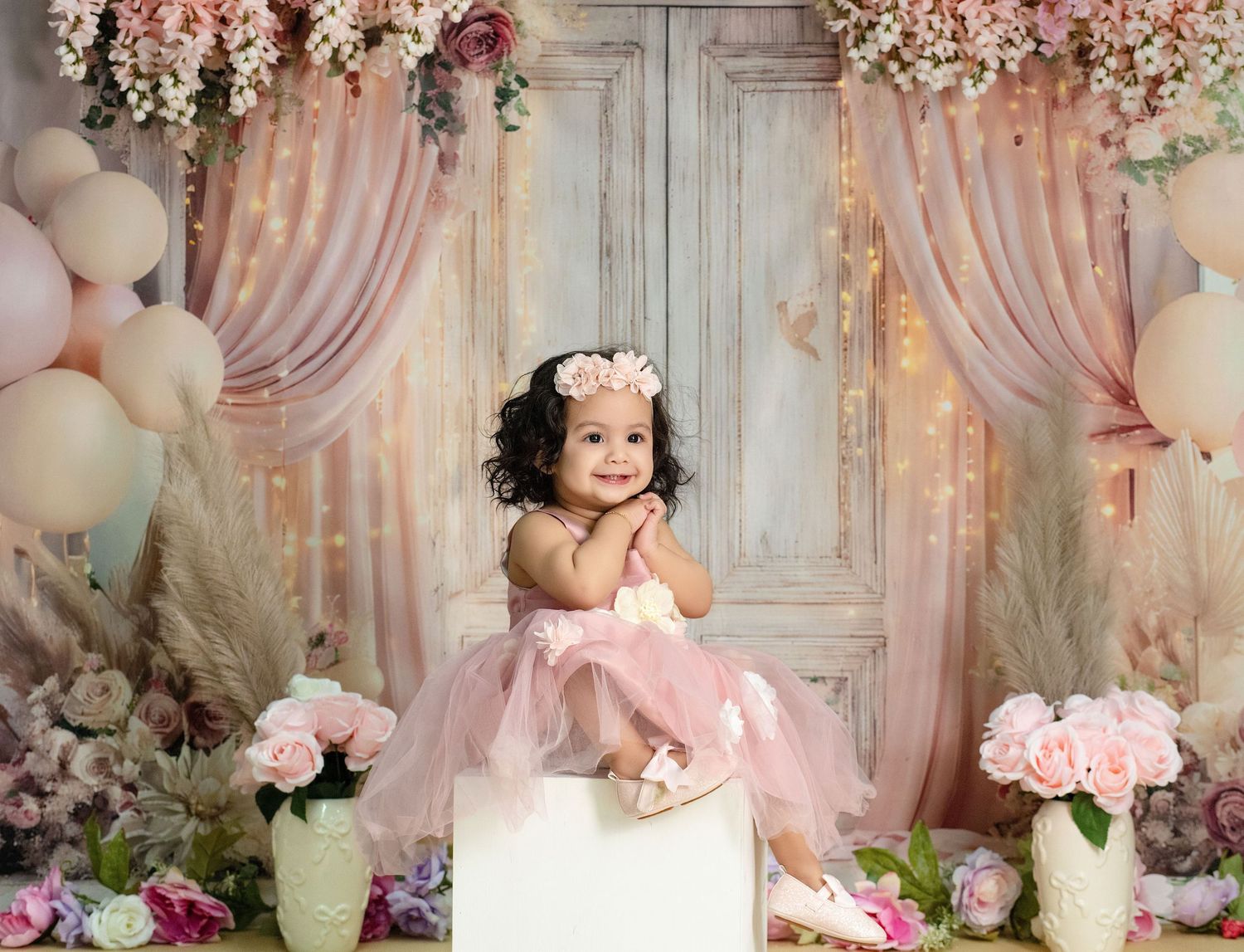 Adorable toddler girl in pink tutu dress sitting among floral decor and balloons in a dreamy studio portrait.