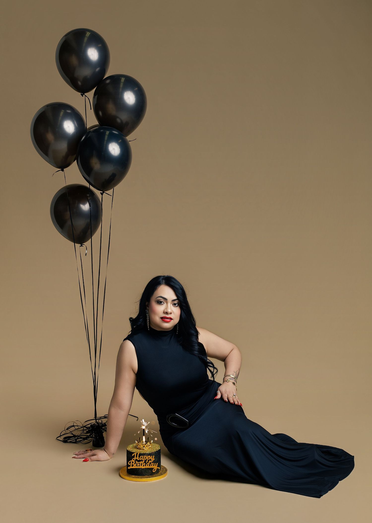 Woman in black gown poses with black balloons and birthday cake on beige background.