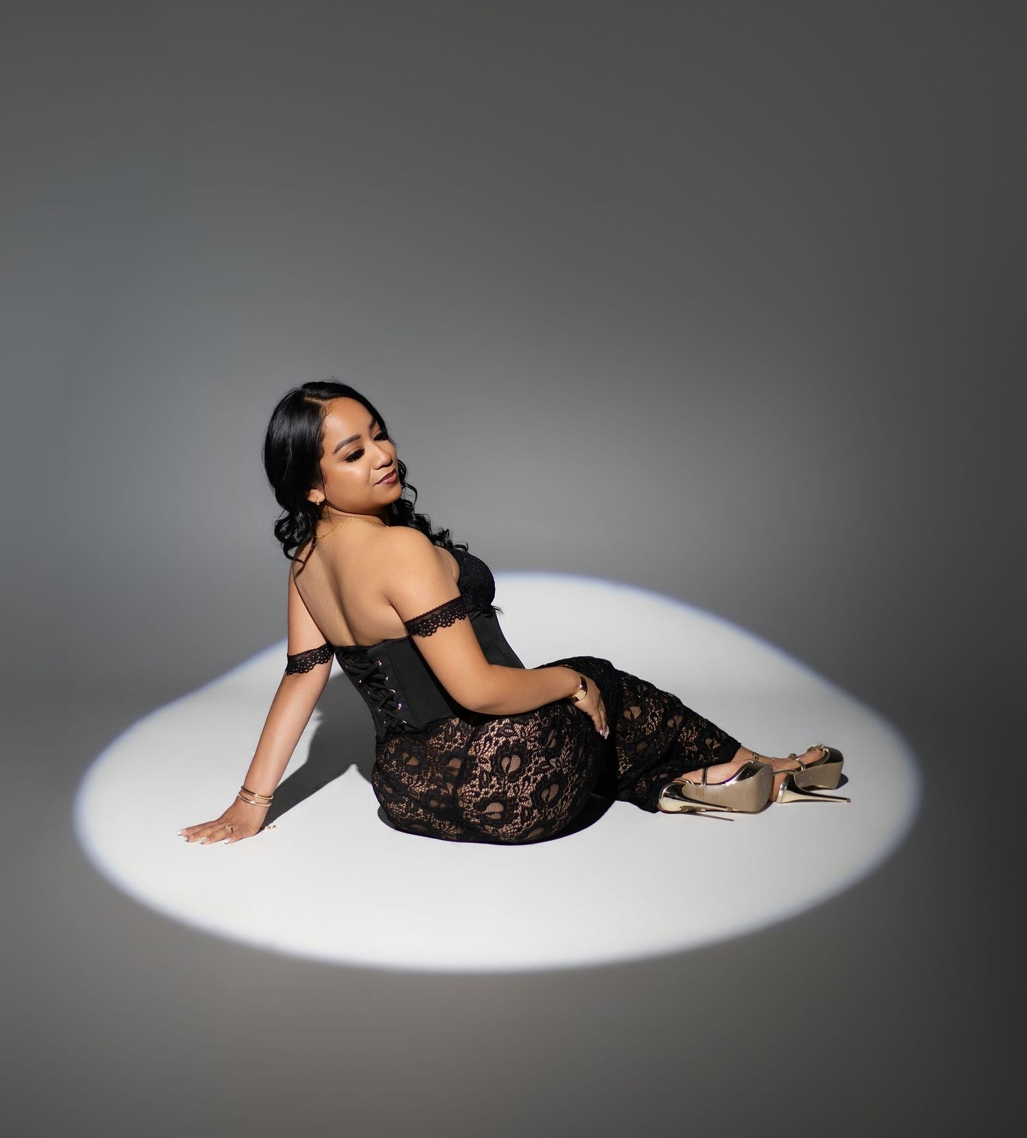 Woman in black lace off-shoulder gown posed elegantly on floor in dramatic spotlight against gray background.