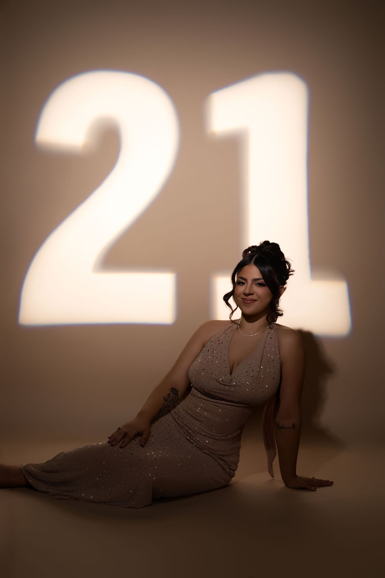 Woman in sparkly beige gown poses on floor before glowing 21 projection for 21st birthday portrait.