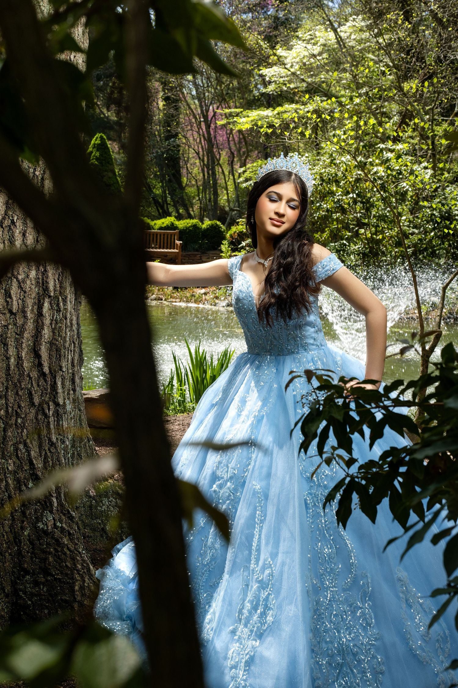 Young woman in a light blue ball gown and tiara poses gracefully in a lush garden near a pond.