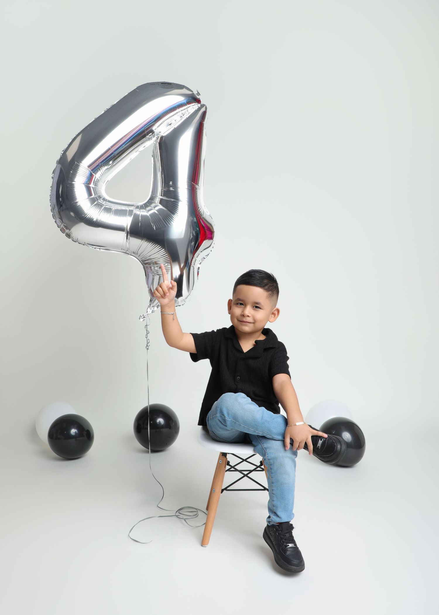 A young boy sits on a stool holding a silver number 4 balloon, wearing a black shirt and jeans, surrounded by black balloons.