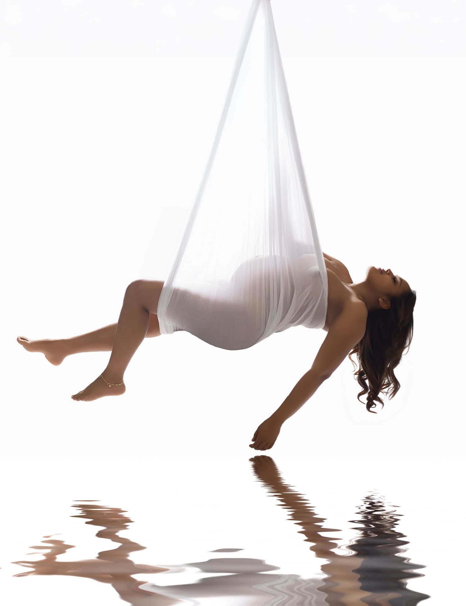 Woman suspended in white aerial silk fabric, gracefully posed above water reflection on white background.