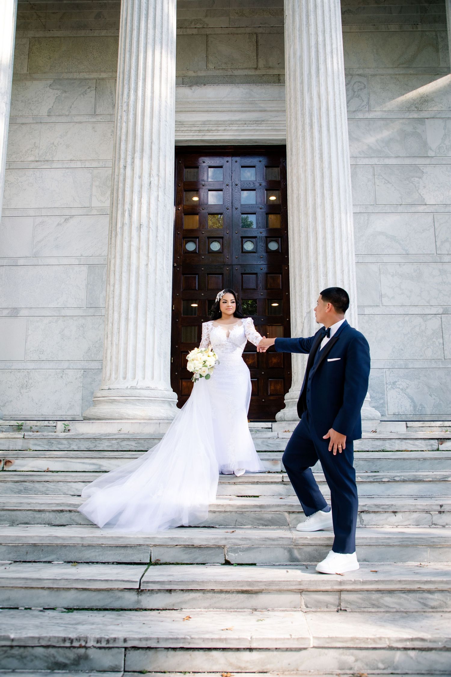 Bride in white gown and groom in navy suit hold hands on marble steps outside elegant building with columns.