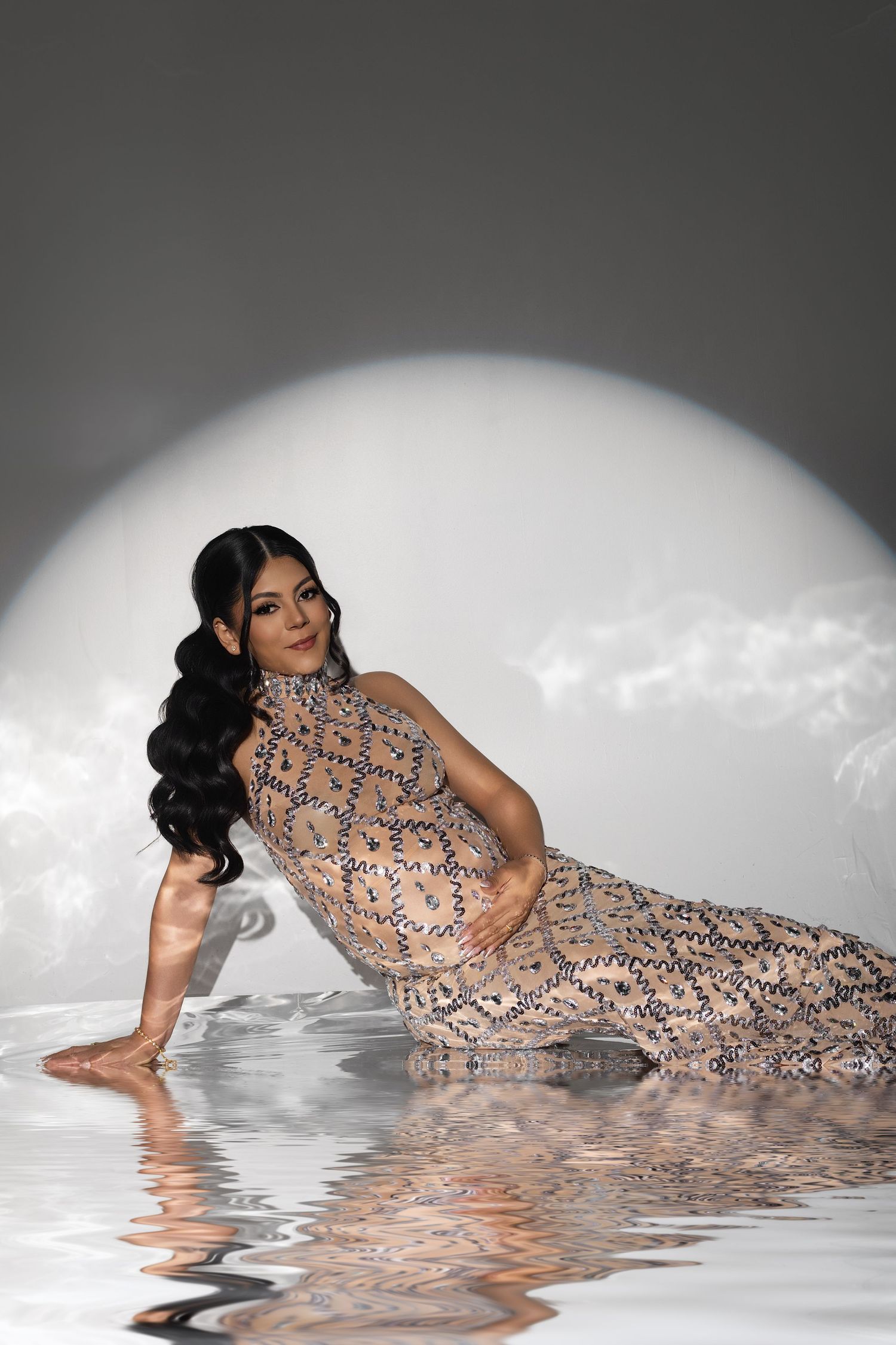 Pregnant woman in sequined gown poses gracefully near reflective water surface against a glowing circular backdrop.
