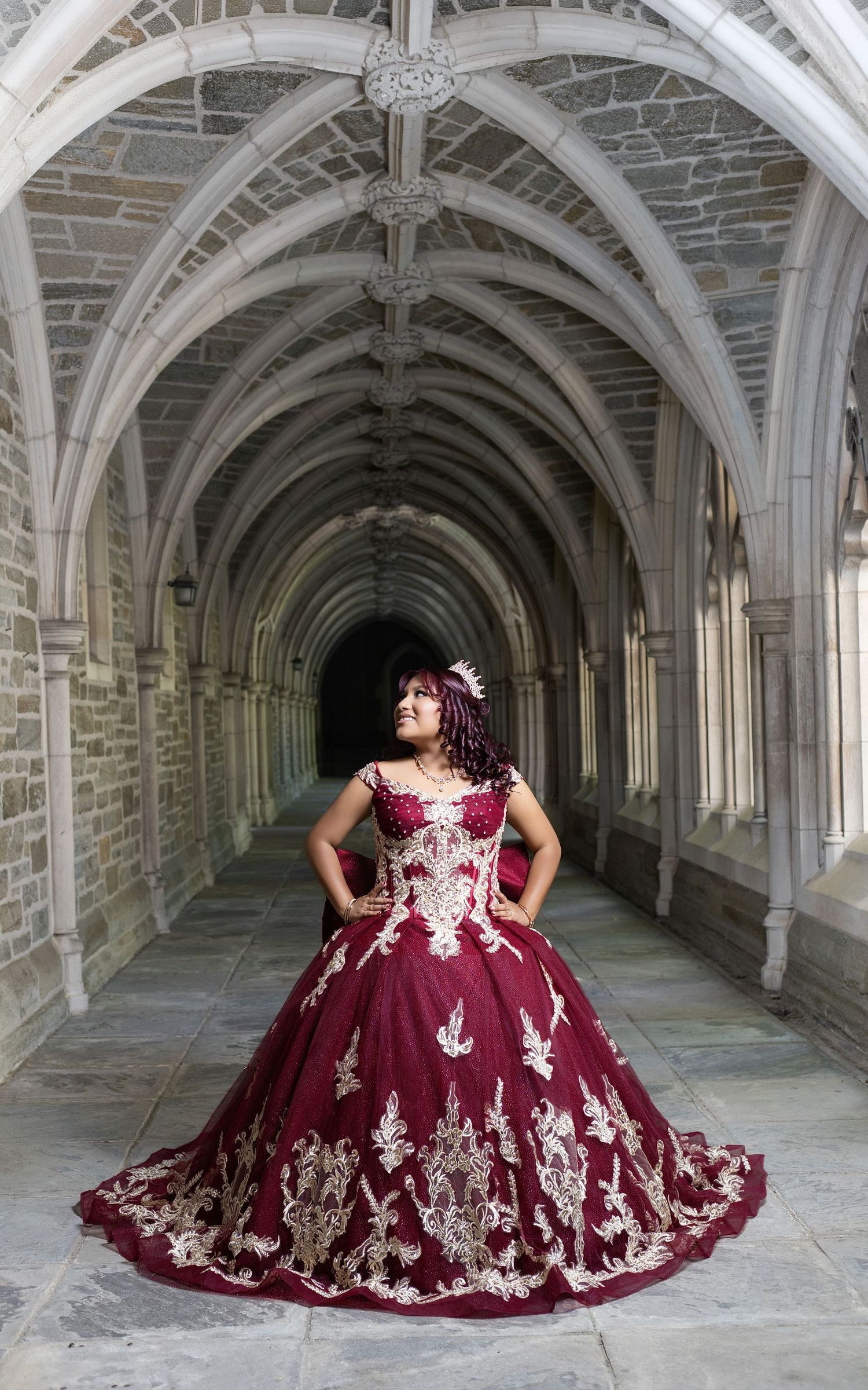 Woman in elegant burgundy and gold ballgown poses in Gothic stone archway corridor.
