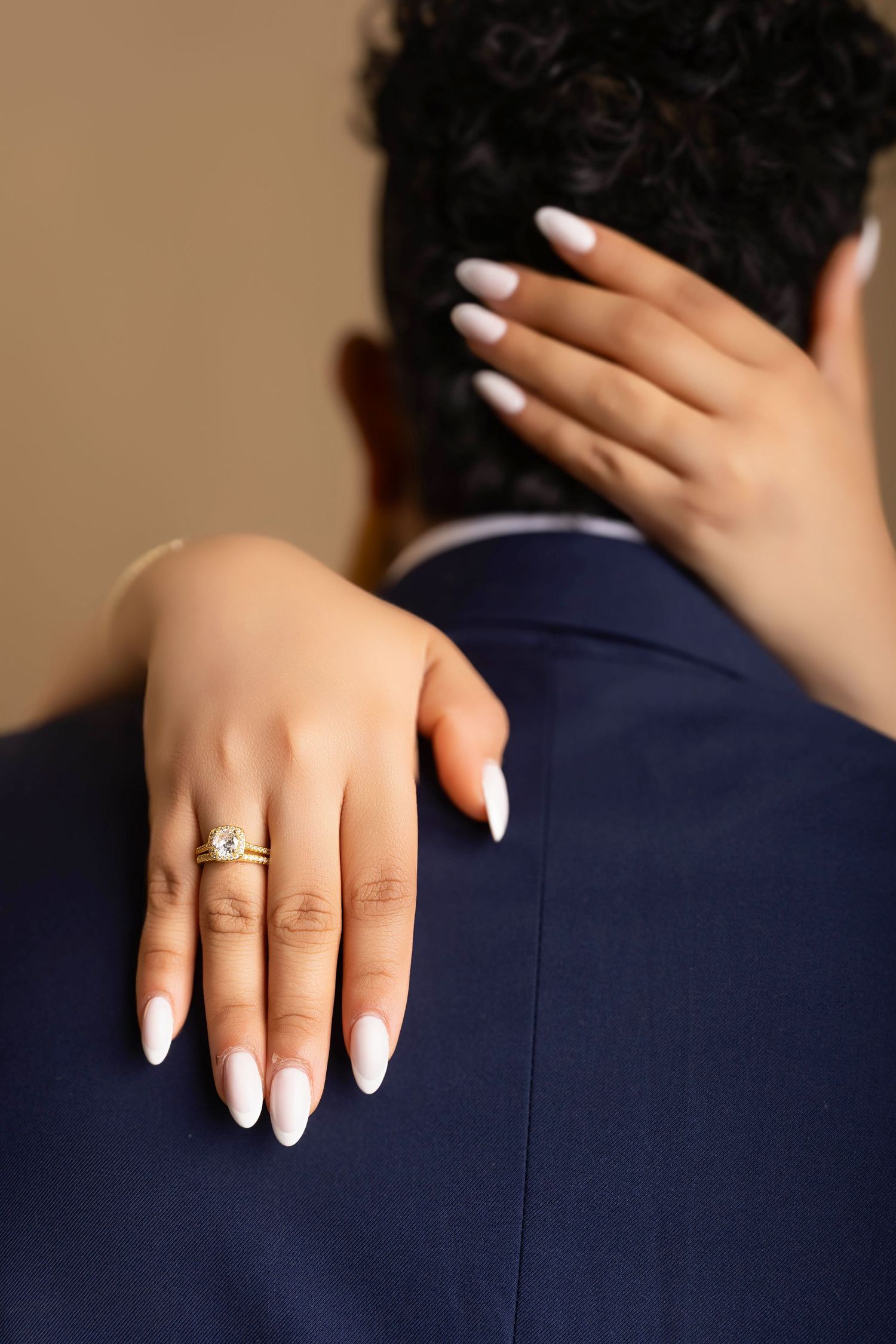 Close-up of a woman's hand with white almond nails and a gold diamond engagement ring resting on a man's navy suit.