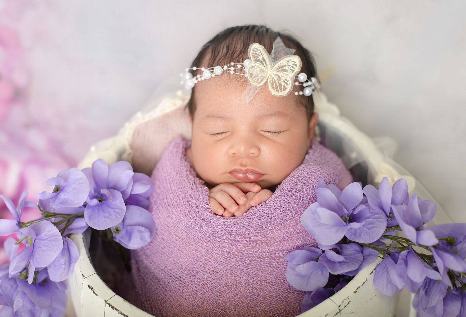 Sleeping newborn baby wrapped in purple swaddle with butterfly headband in white basket surrounded by lavender flowers.
