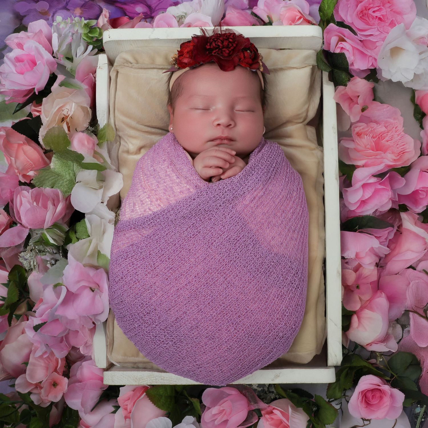 Newborn baby wrapped in purple swaddle with red floral headband sleeping in white wooden box surrounded by pink roses.