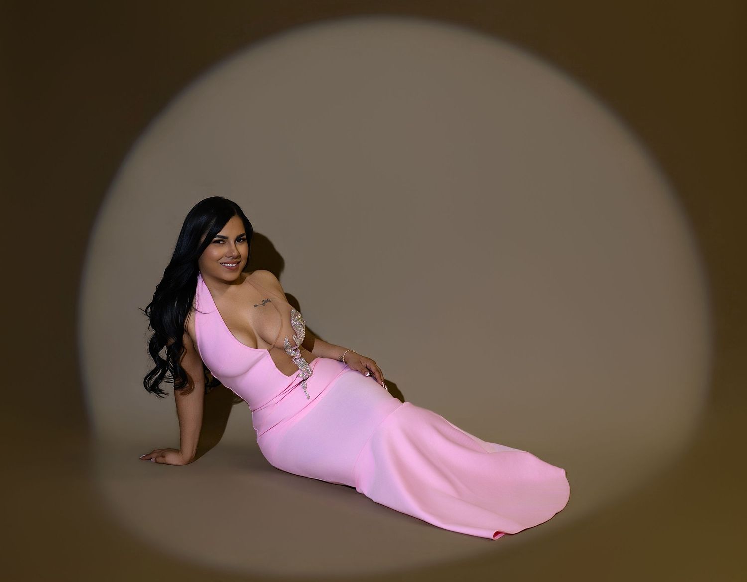 Woman in pink one-shoulder gown poses elegantly on floor in spotlight against warm brown background.