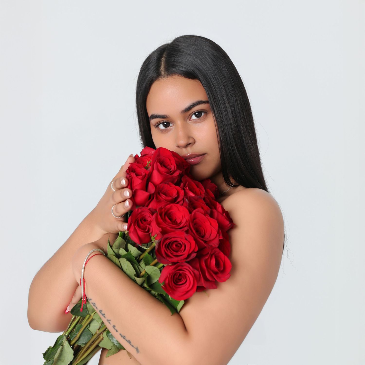 Young woman with long dark hair holding a large bouquet of red roses, with visible tattoos on her arm.