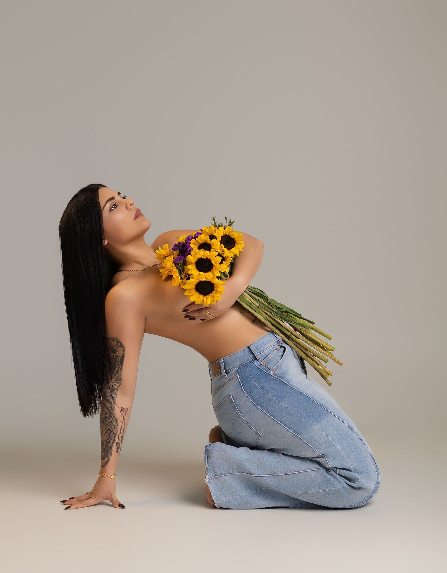 Woman kneeling with long black hair, tattoo, holding sunflowers against chest, wearing blue jeans, on gray background.