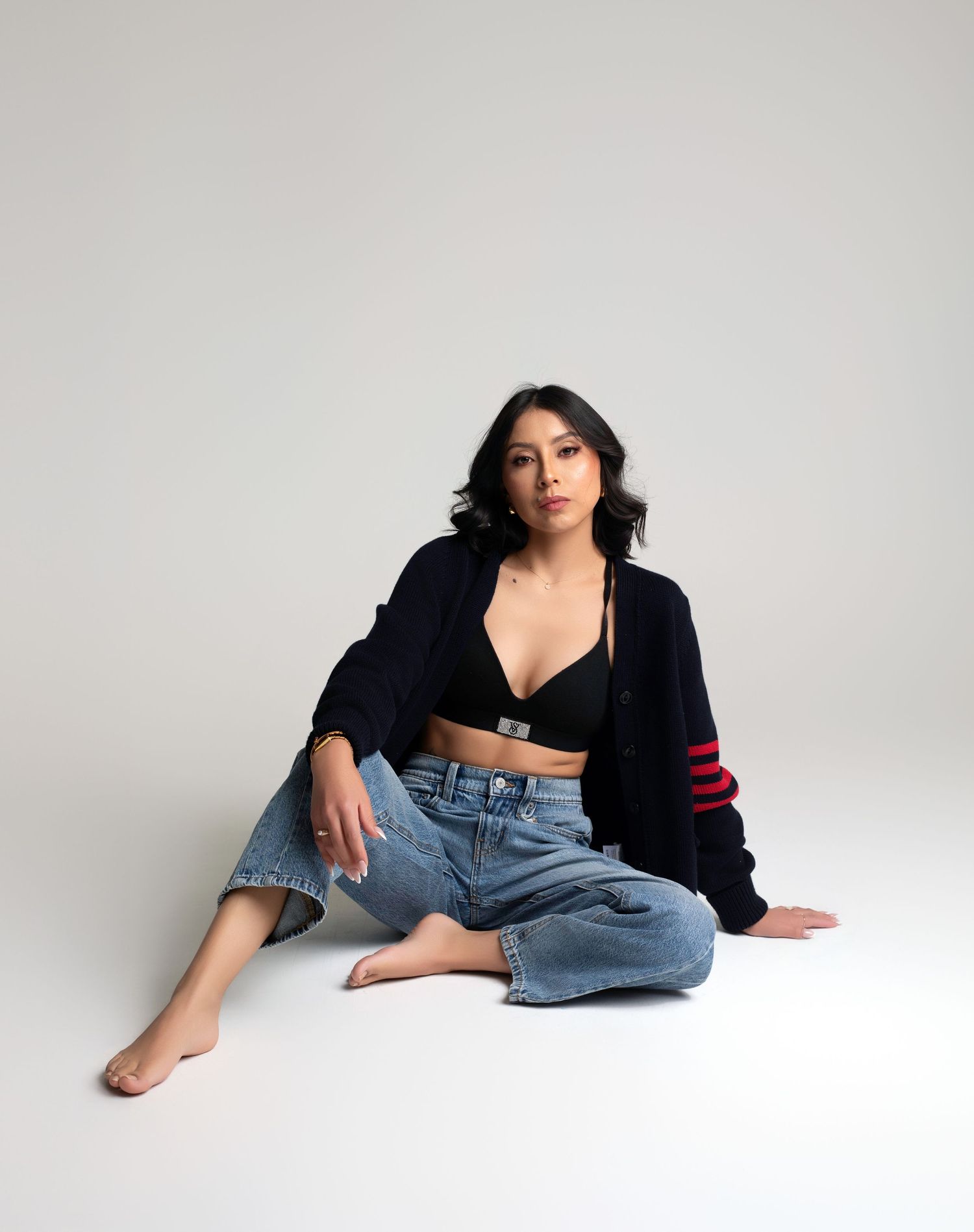Woman in black cardigan, crop top, and jeans sitting on white floor in casual fashion pose.