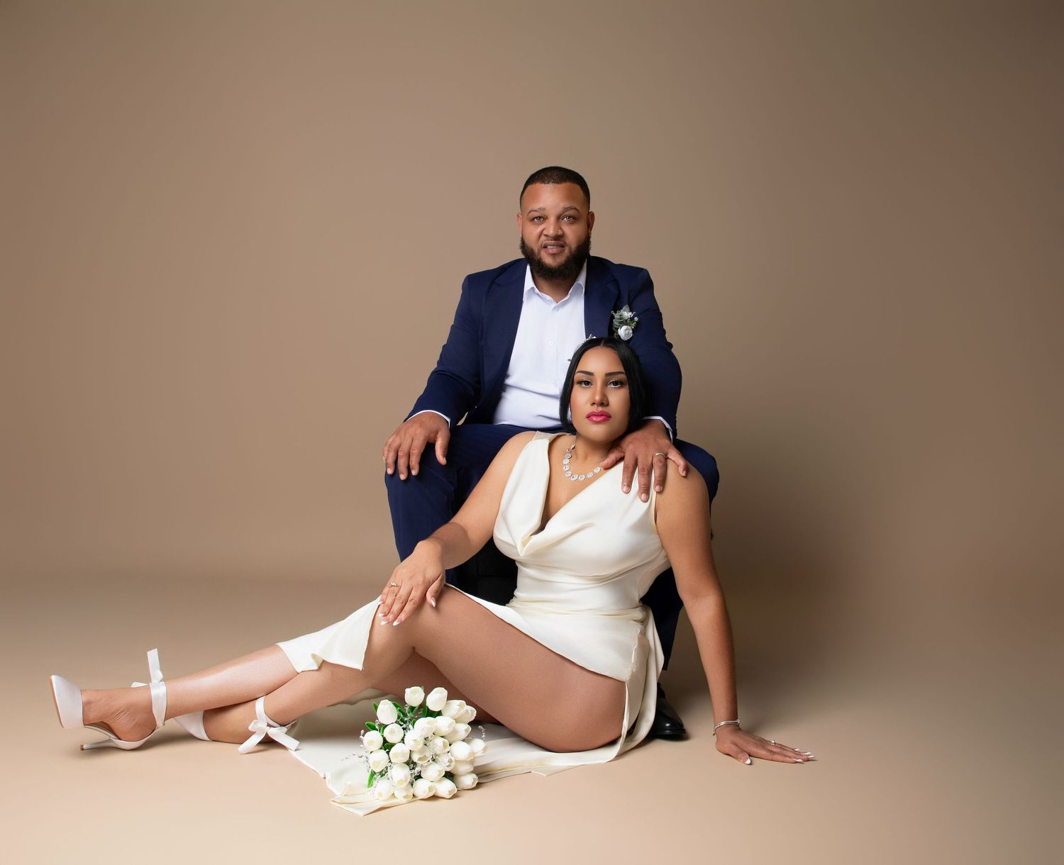 Couple in wedding attire posing in studio, man in blue suit seated behind woman in white dress holding flowers.