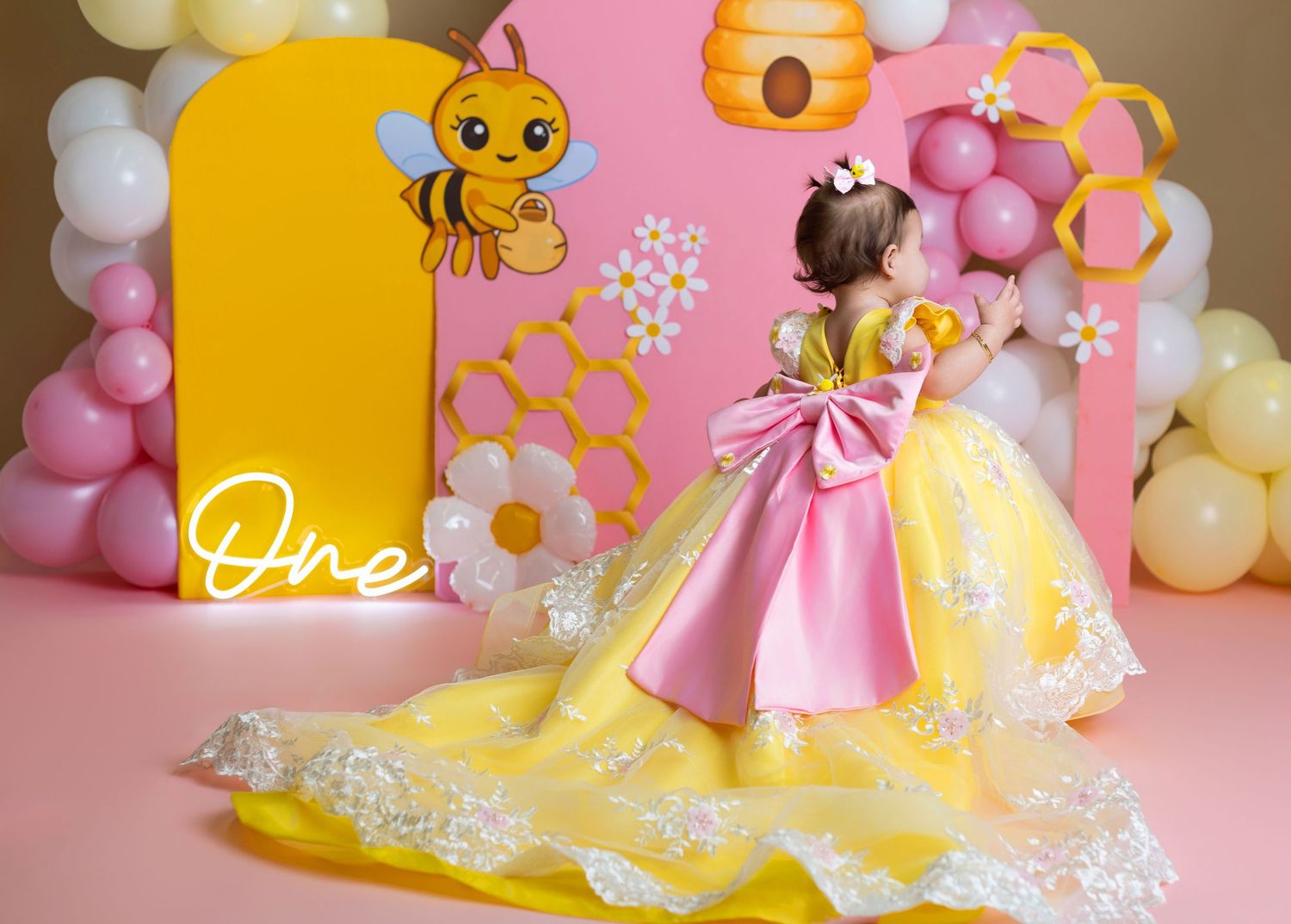 A baby girl in a yellow and pink floral dress with a bow poses at a bee-themed first birthday party.