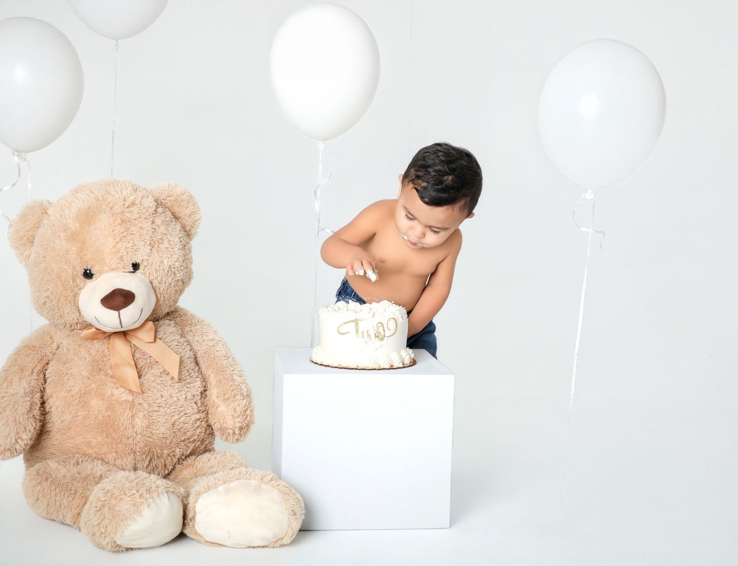 Toddler boy smashing a second birthday cake beside a large teddy bear, surrounded by white balloons.