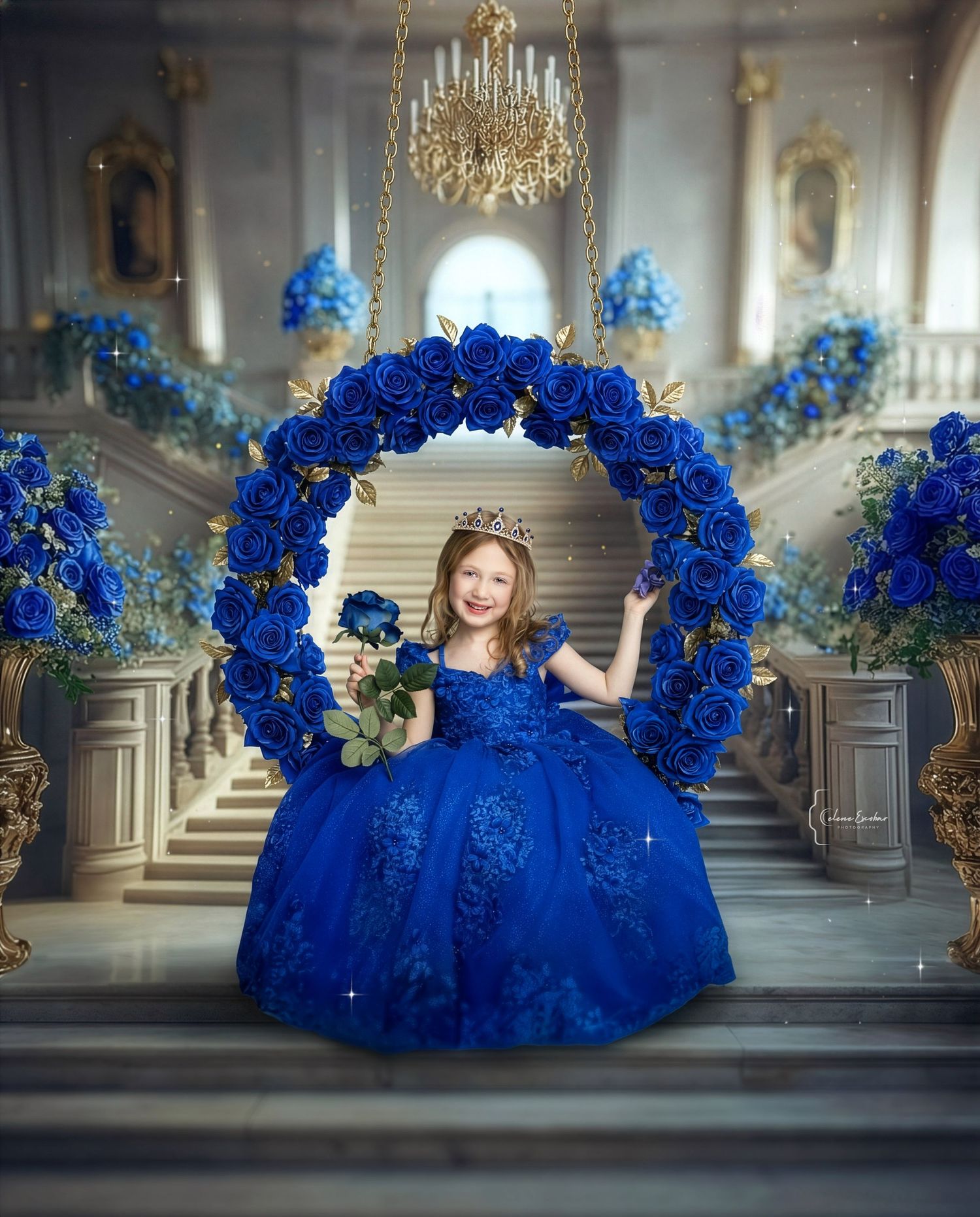 Young girl in royal blue ball gown with crown sits on floral swing surrounded by blue roses in elegant ballroom.