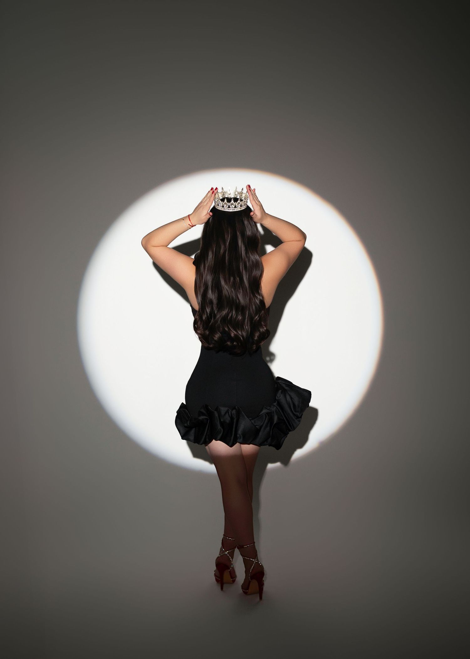 Woman in black dress and heels placing a crown on her head, posed in a dramatic spotlight from behind.