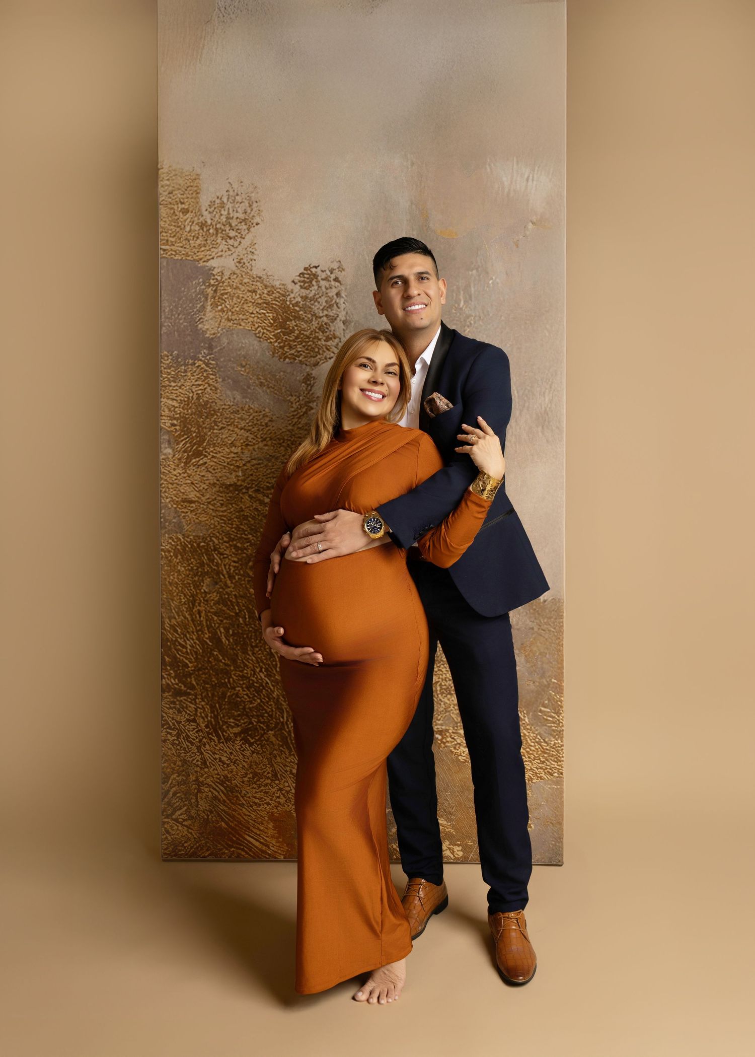 Couple maternity photo, woman in rust orange gown cradling bump, man in navy suit embracing her, studio backdrop.