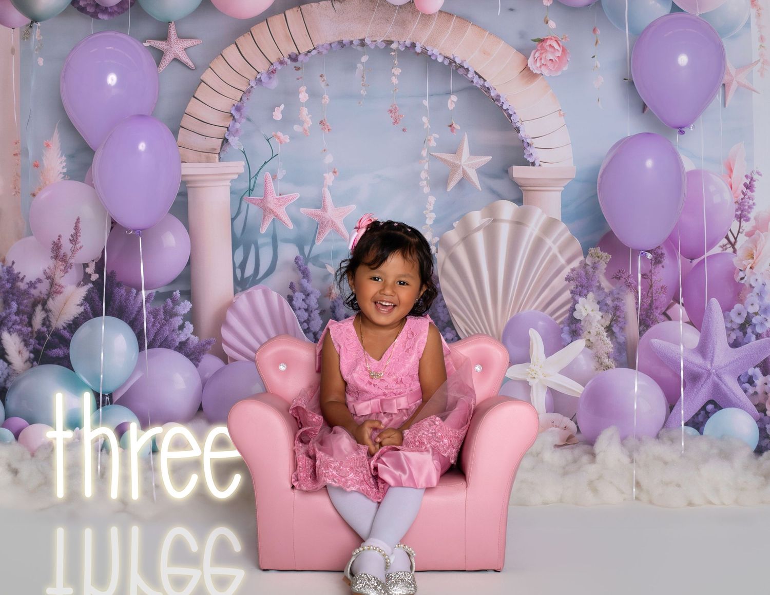 A toddler girl sits in a pink chair surrounded by purple balloons and seashells at a mermaid-themed third birthday party.