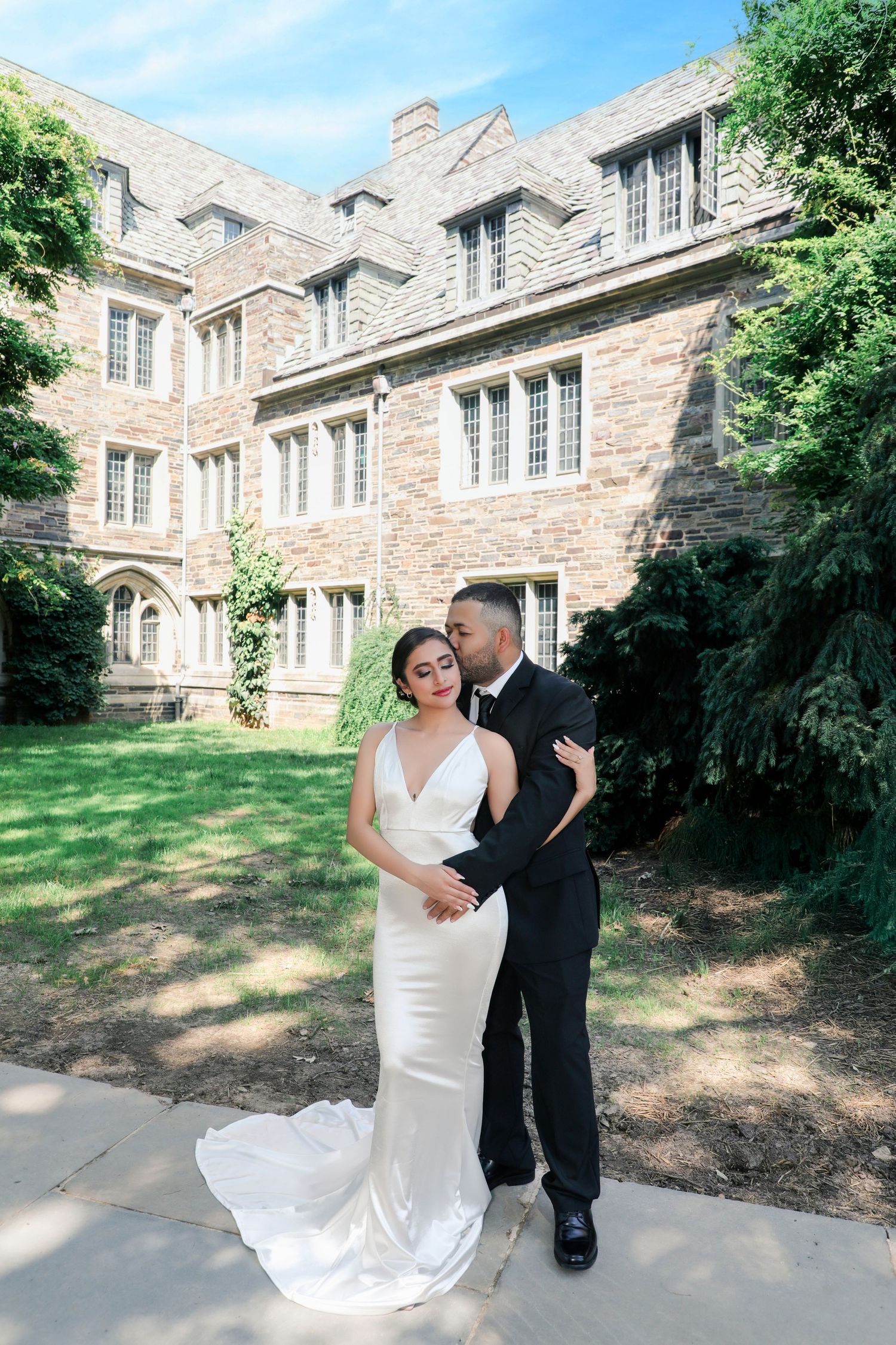 Groom in black tux kisses bride in sleek white gown outside elegant stone building.