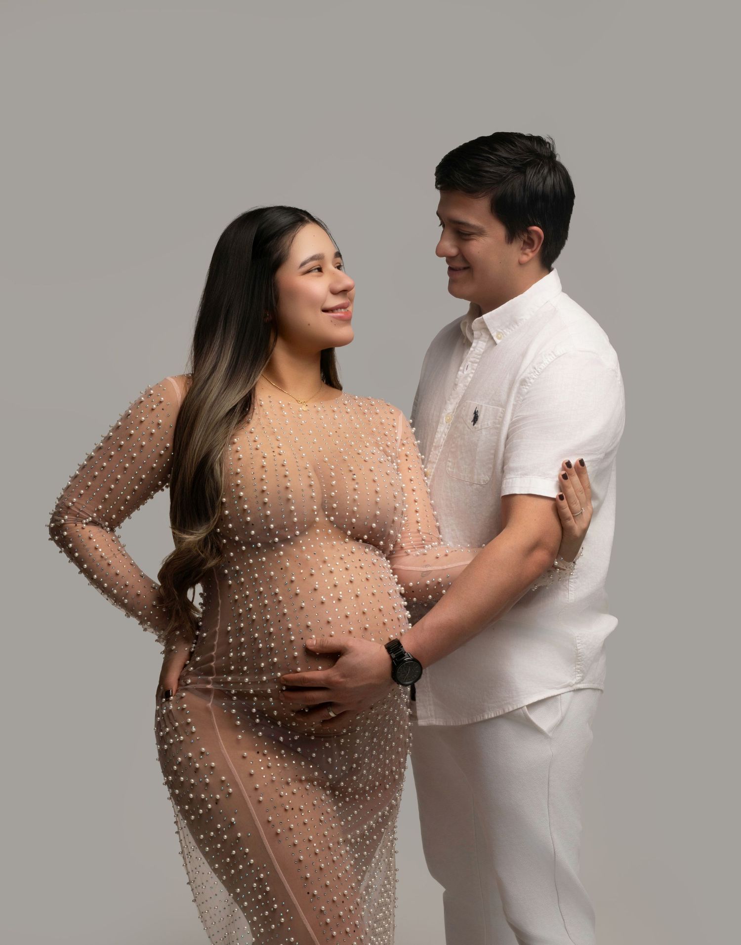 Couple in maternity photoshoot, woman in pearl mesh gown cradling baby bump, man in white outfit embracing her.
