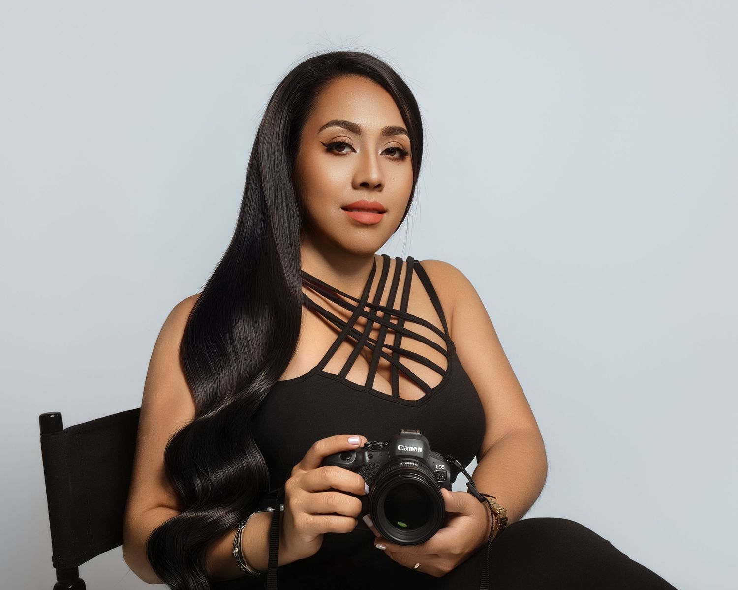 Woman in black outfit holding a DSLR camera, seated against a light gray background, with long dark hair.