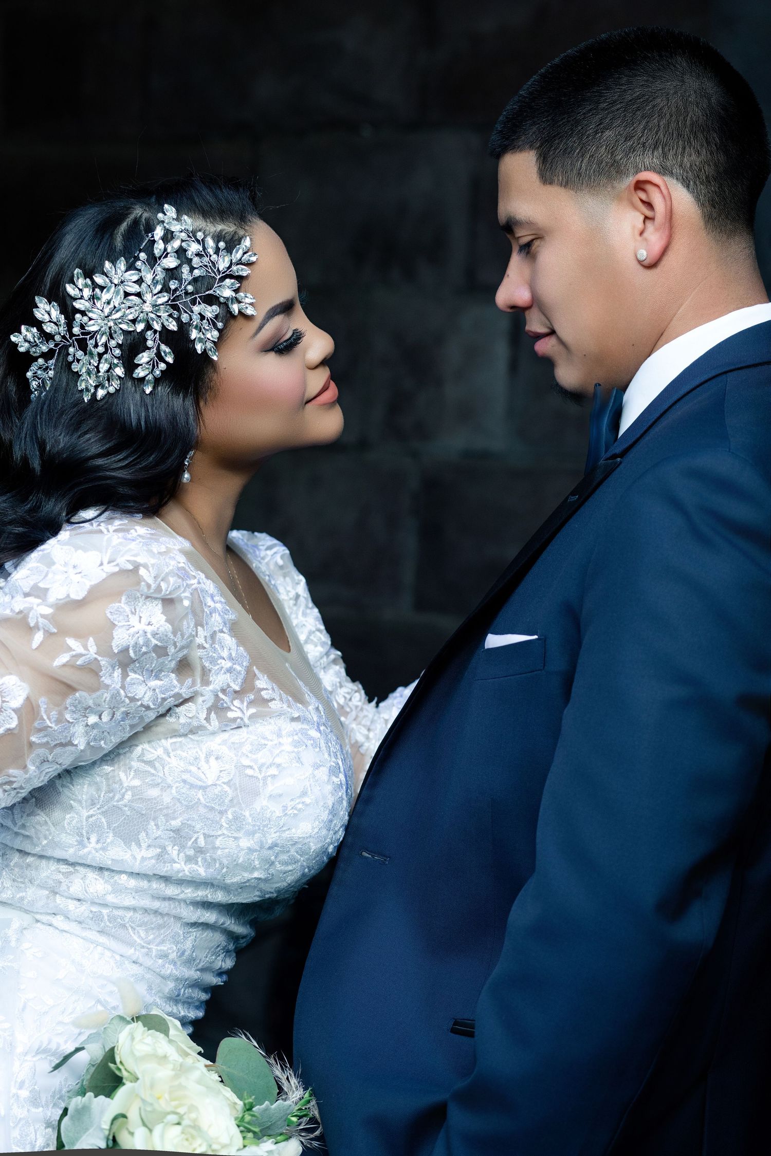 A bride and groom in elegant wedding attire share an intimate moment, she wearing a lace gown and crystal hair piece.