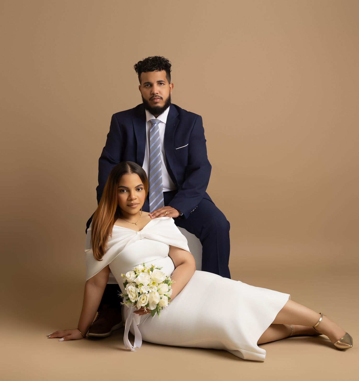 Elegant couple in wedding attire, groom in navy suit, bride in white dress holding bouquet, posed on beige background.