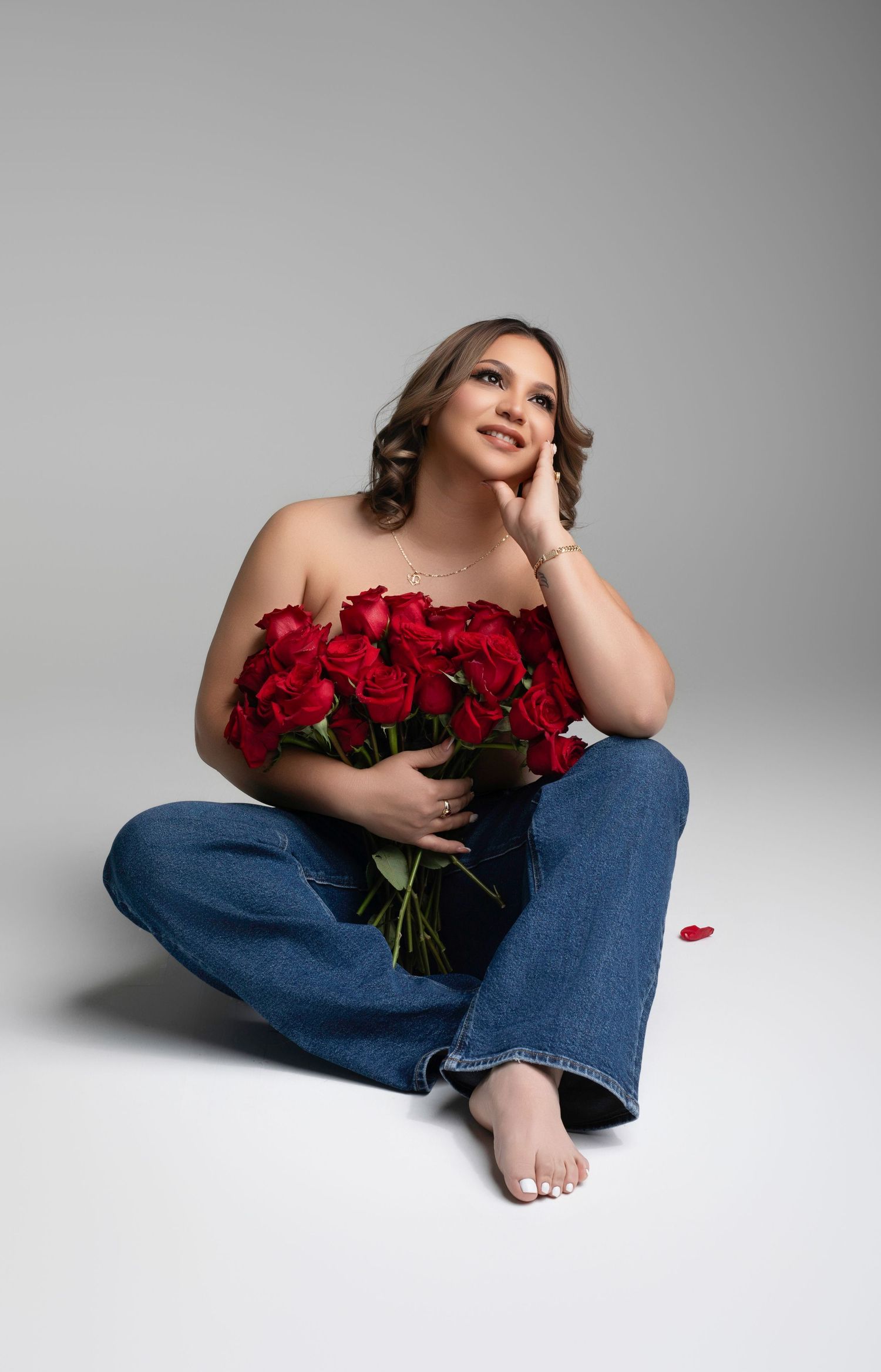 Woman sitting on floor holding red roses bouquet, wearing blue jeans and strapless top, looking up dreamily.