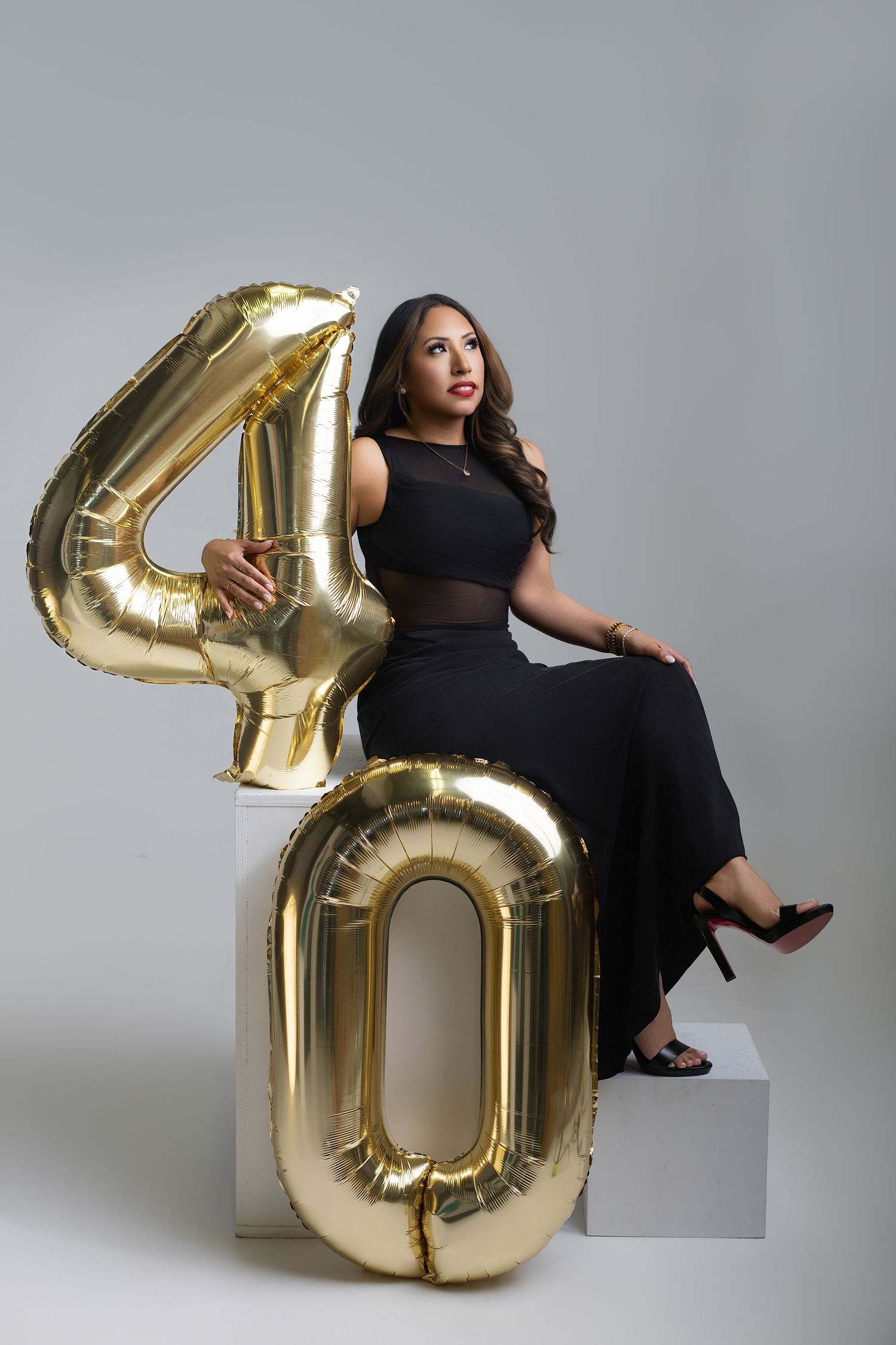 Woman in black dress sitting with gold number 40 balloons, celebrating her 40th birthday in a studio.
