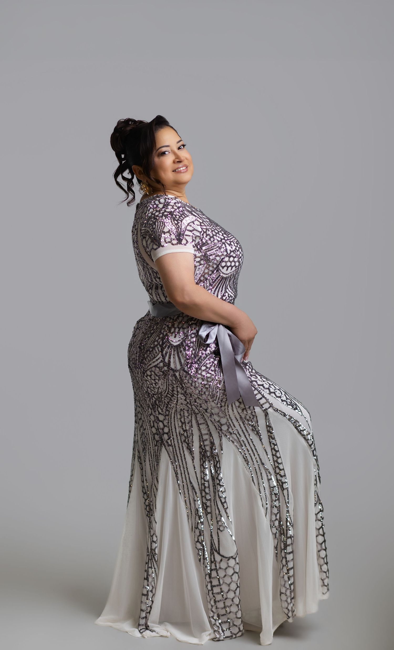 Woman in elegant purple and black lace mermaid gown with white sash, posing gracefully in studio setting.