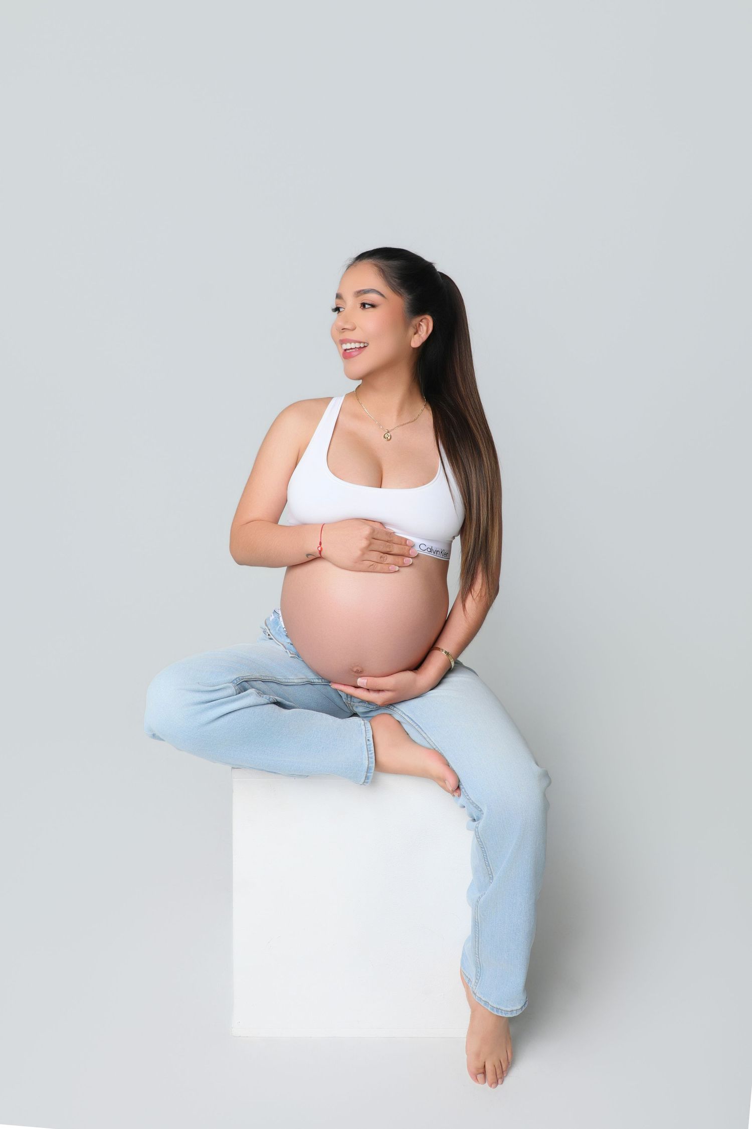 Smiling pregnant woman in white crop top and jeans sitting on white cube, cradling baby bump against gray background.