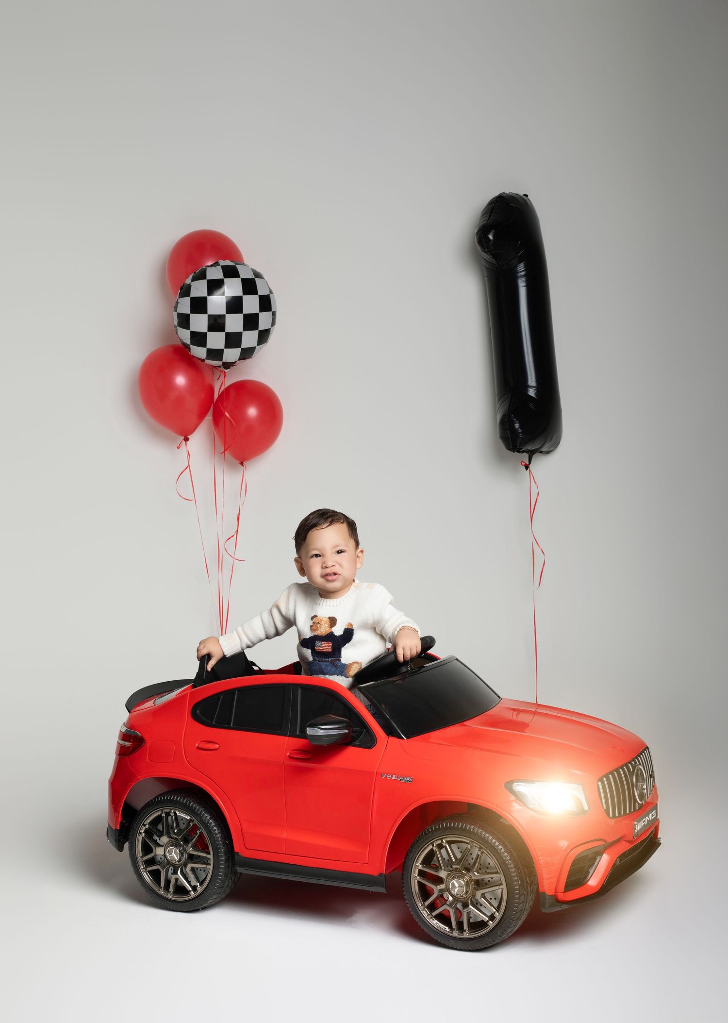 Baby boy sits in red toy Mercedes car with race balloons celebrating his 1st birthday.