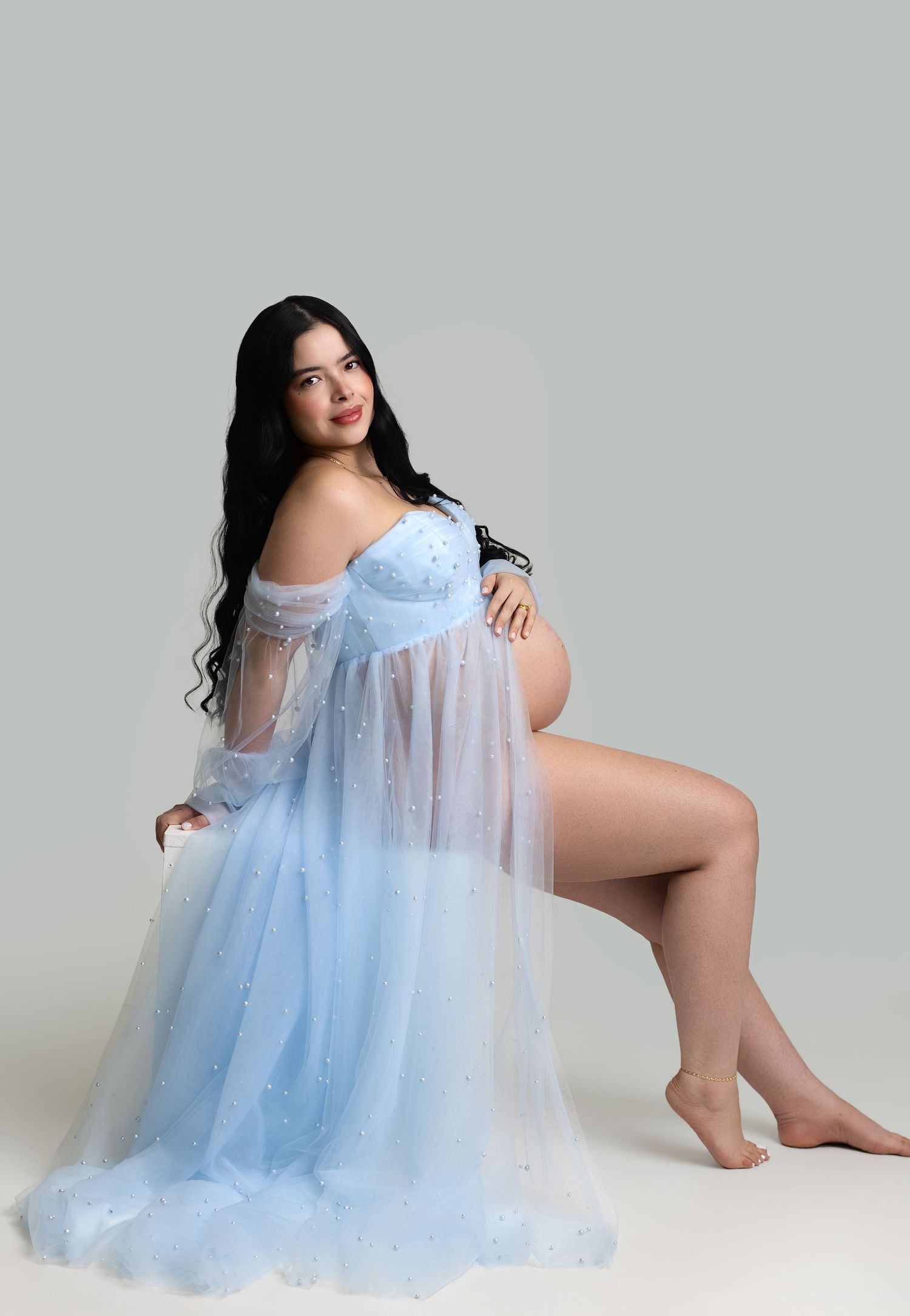 Pregnant woman in sheer light blue maternity gown posing gracefully in a studio photography session.