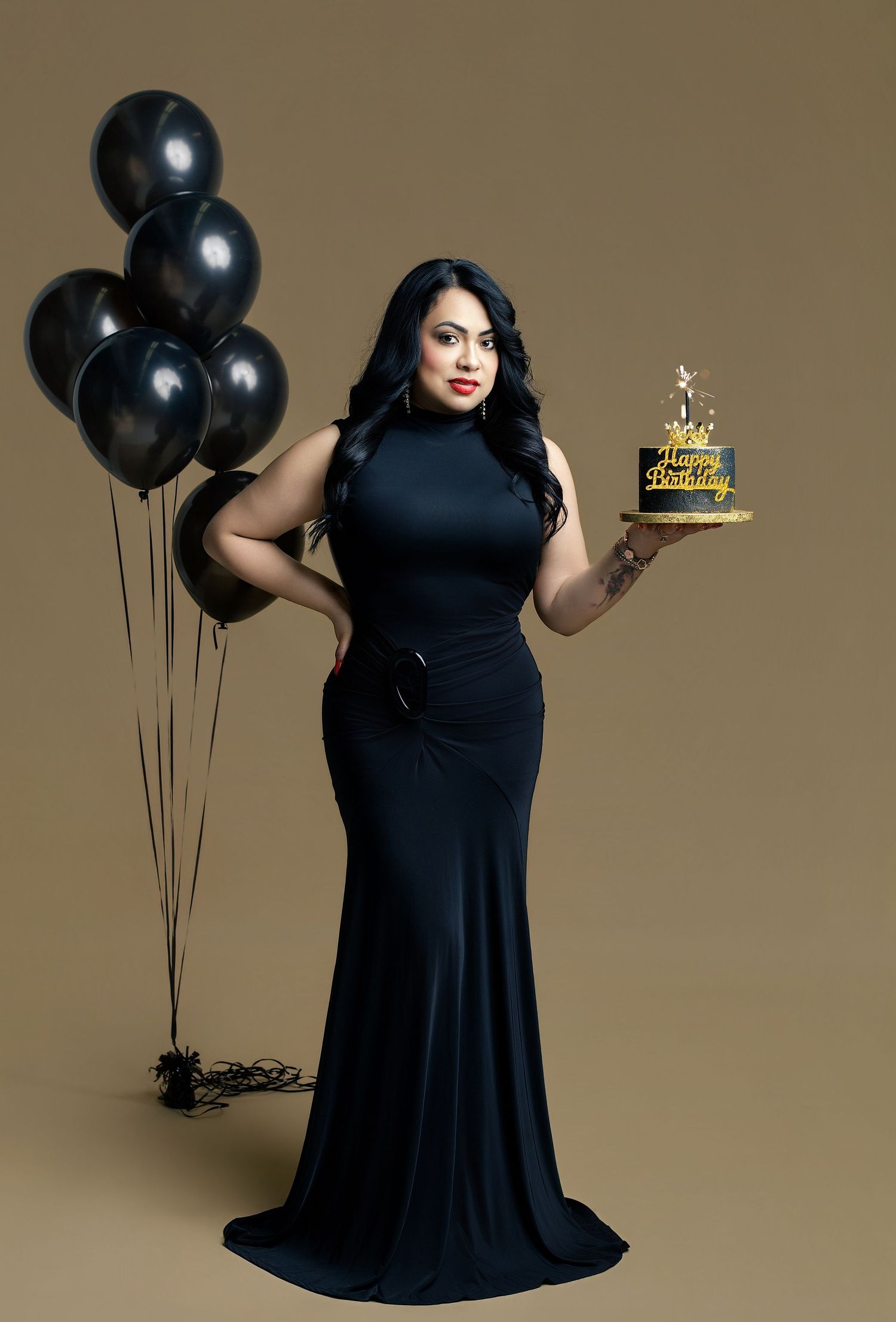 Woman in black gown holding Happy Birthday cake with black balloons on gold background.