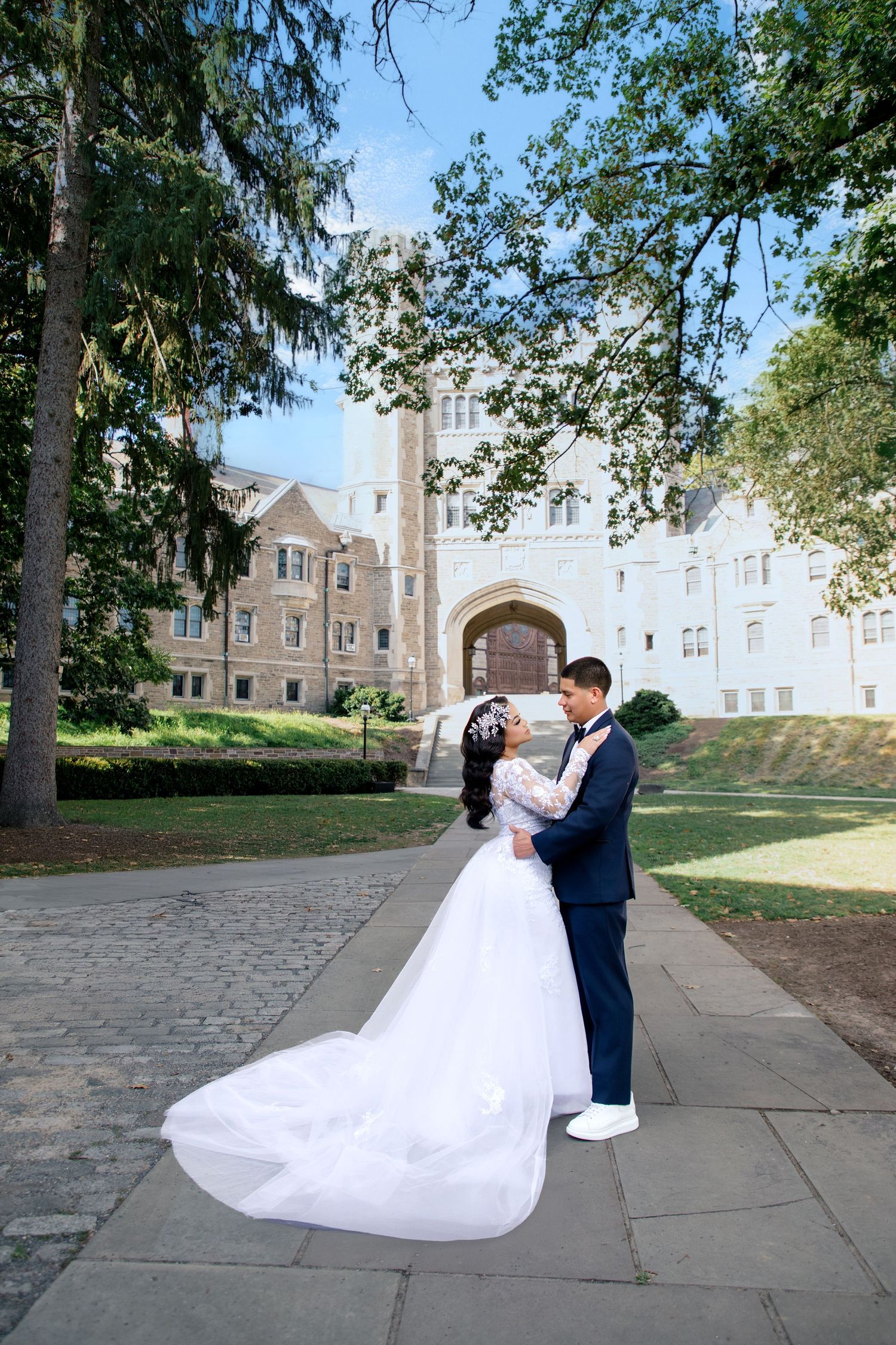 Bride and groom embrace on a stone path outside a historic castle-like building surrounded by lush green trees.