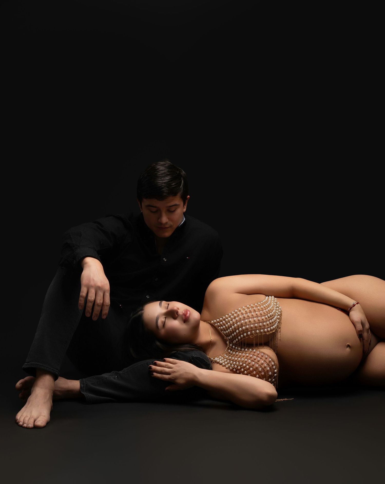 A pregnant woman in a beaded top reclines on the floor beside a man in a black suit against a dark background.