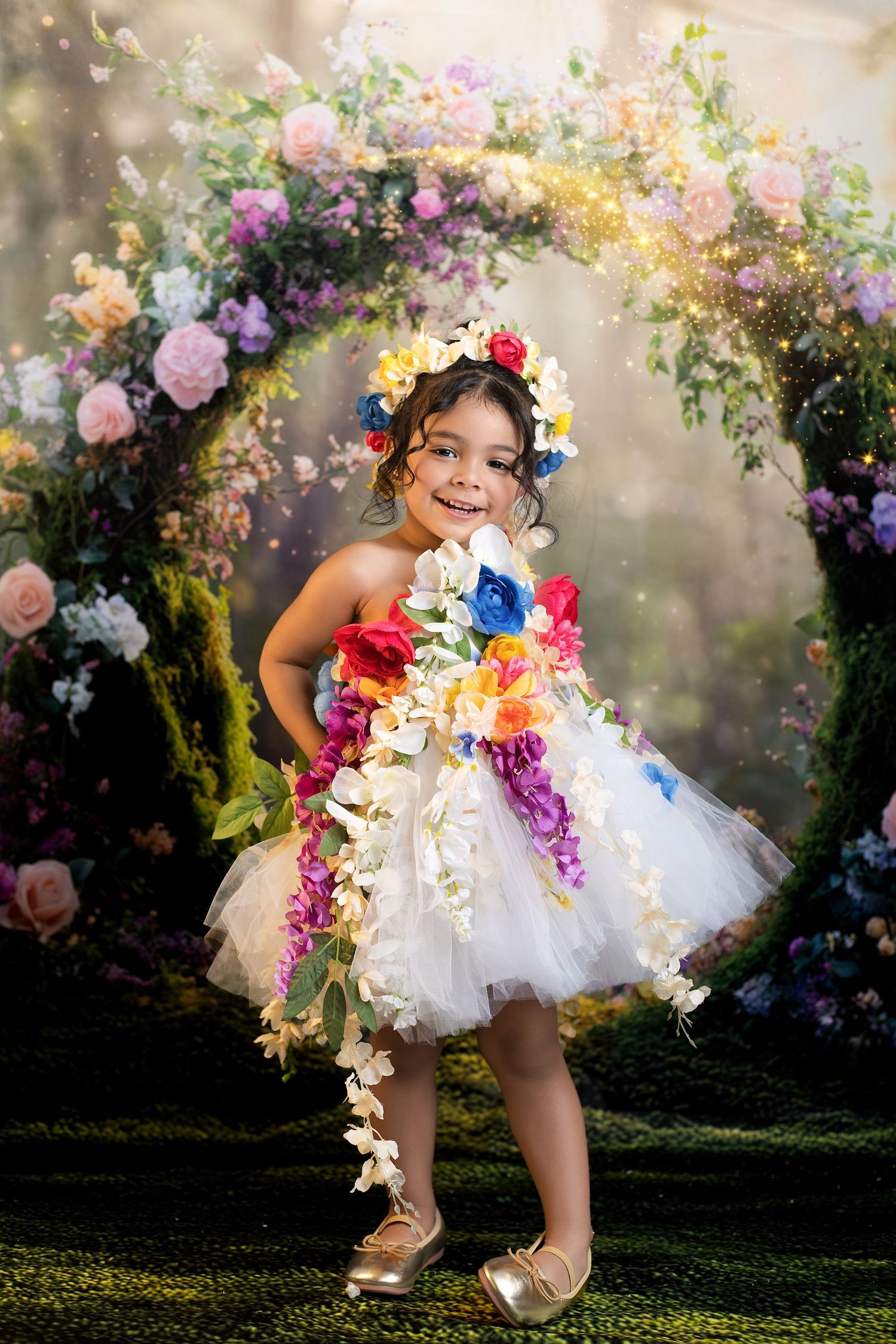Young girl in white tulle dress adorned with colorful flowers poses before a floral arch in enchanted garden setting.