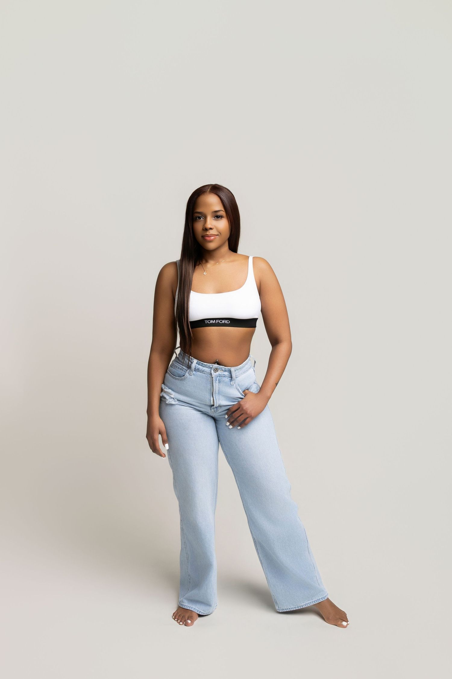 Young woman wearing a white Calvin Klein crop top and light blue wide-leg jeans, posing barefoot on a gray background.