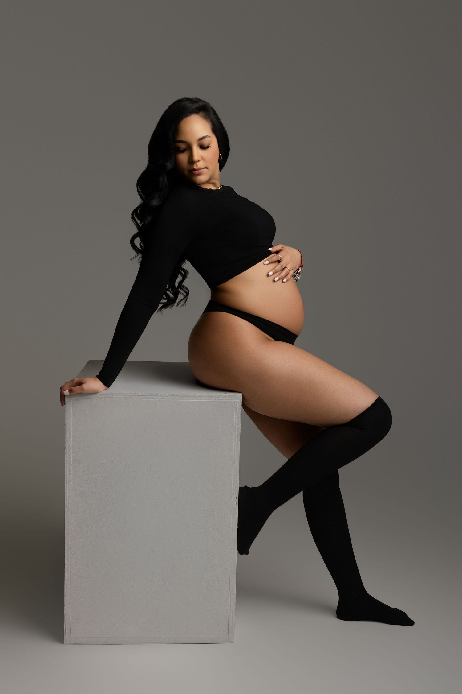Pregnant woman in black crop top, underwear, and knee-high socks posing beside a white cube in a studio.