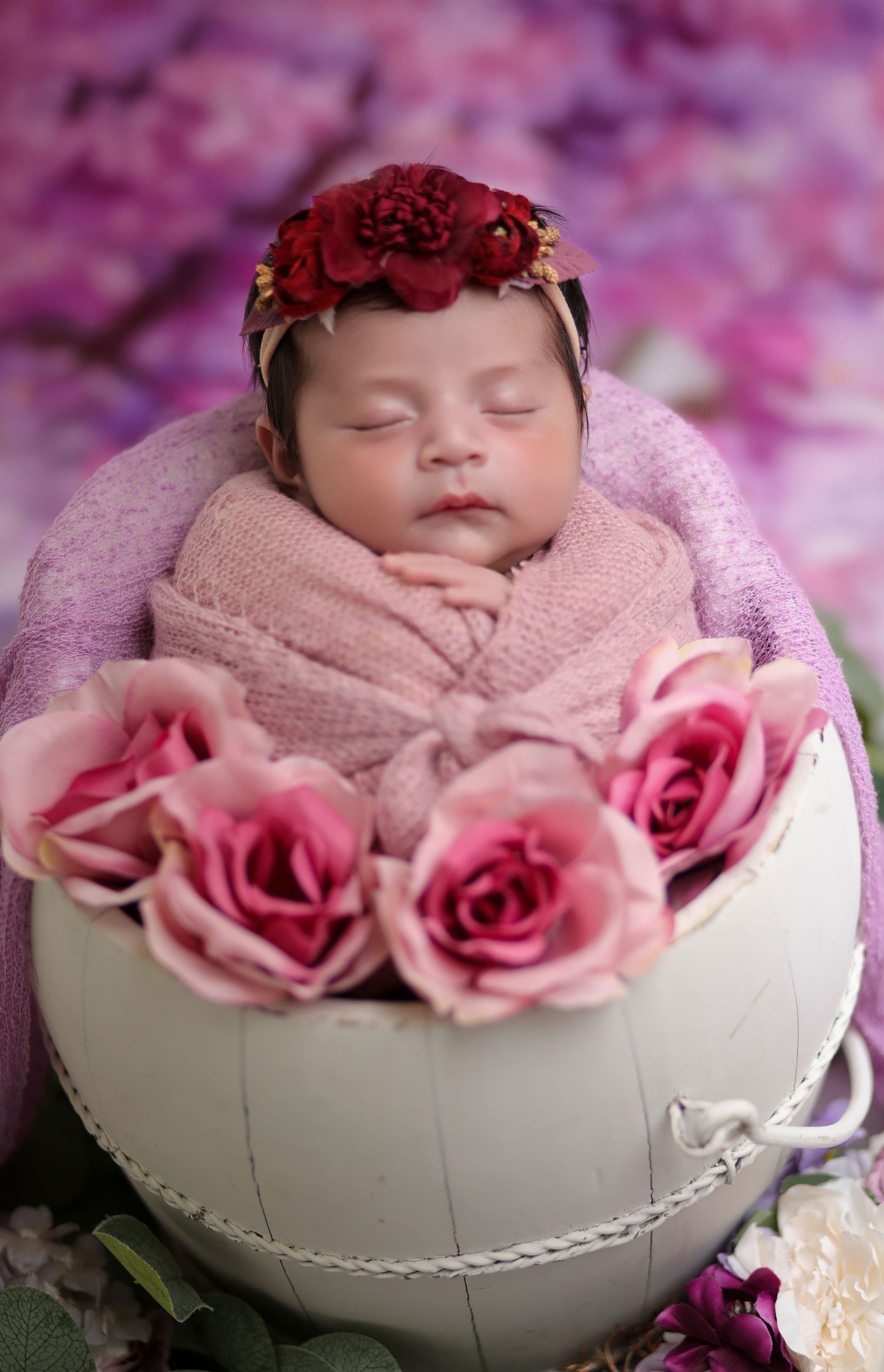 Sleeping newborn baby wrapped in pink, wearing a red floral crown, resting in a white bowl with pink roses.