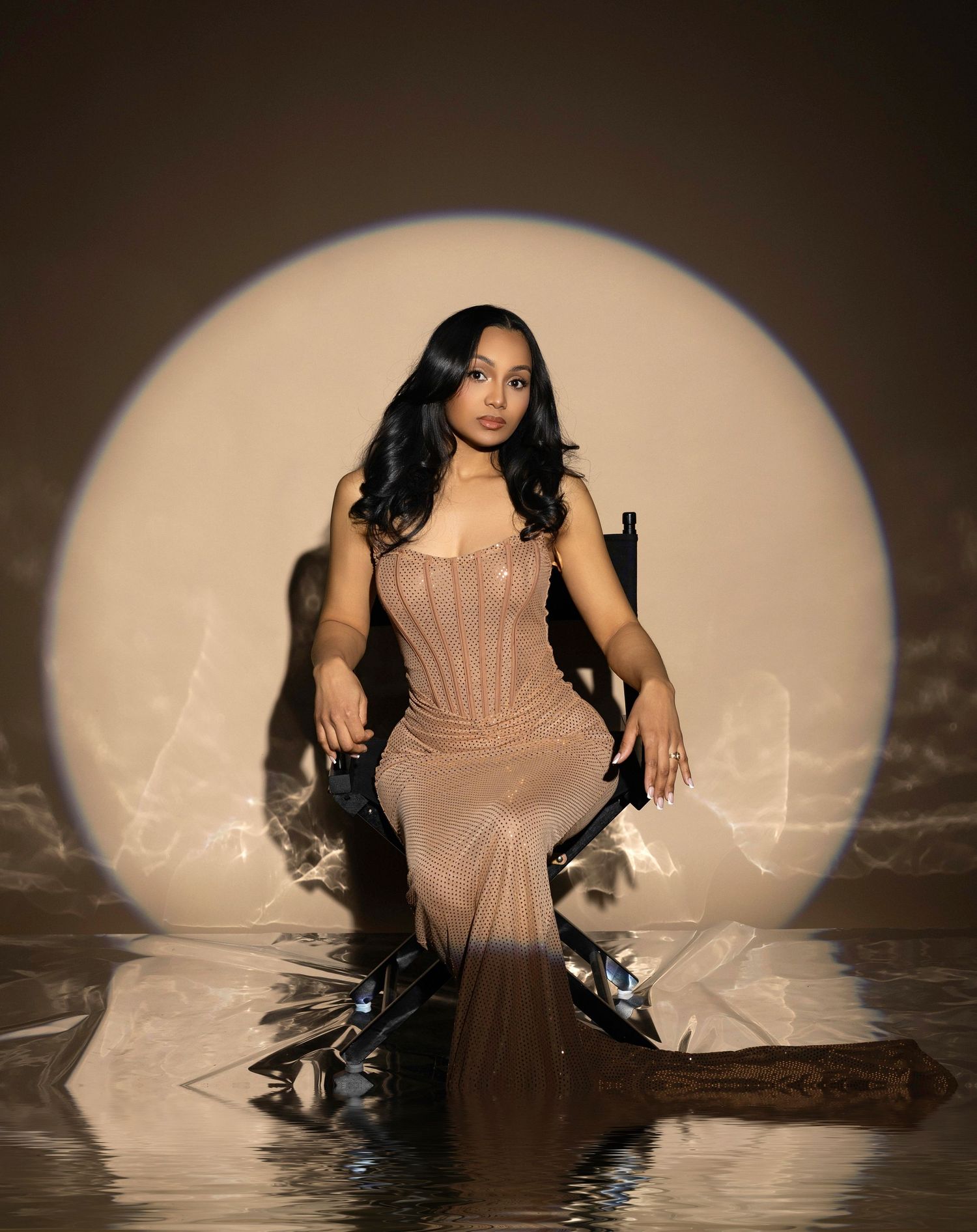 A woman in a sheer nude corset gown sits elegantly before a glowing circular backdrop reflected in water.